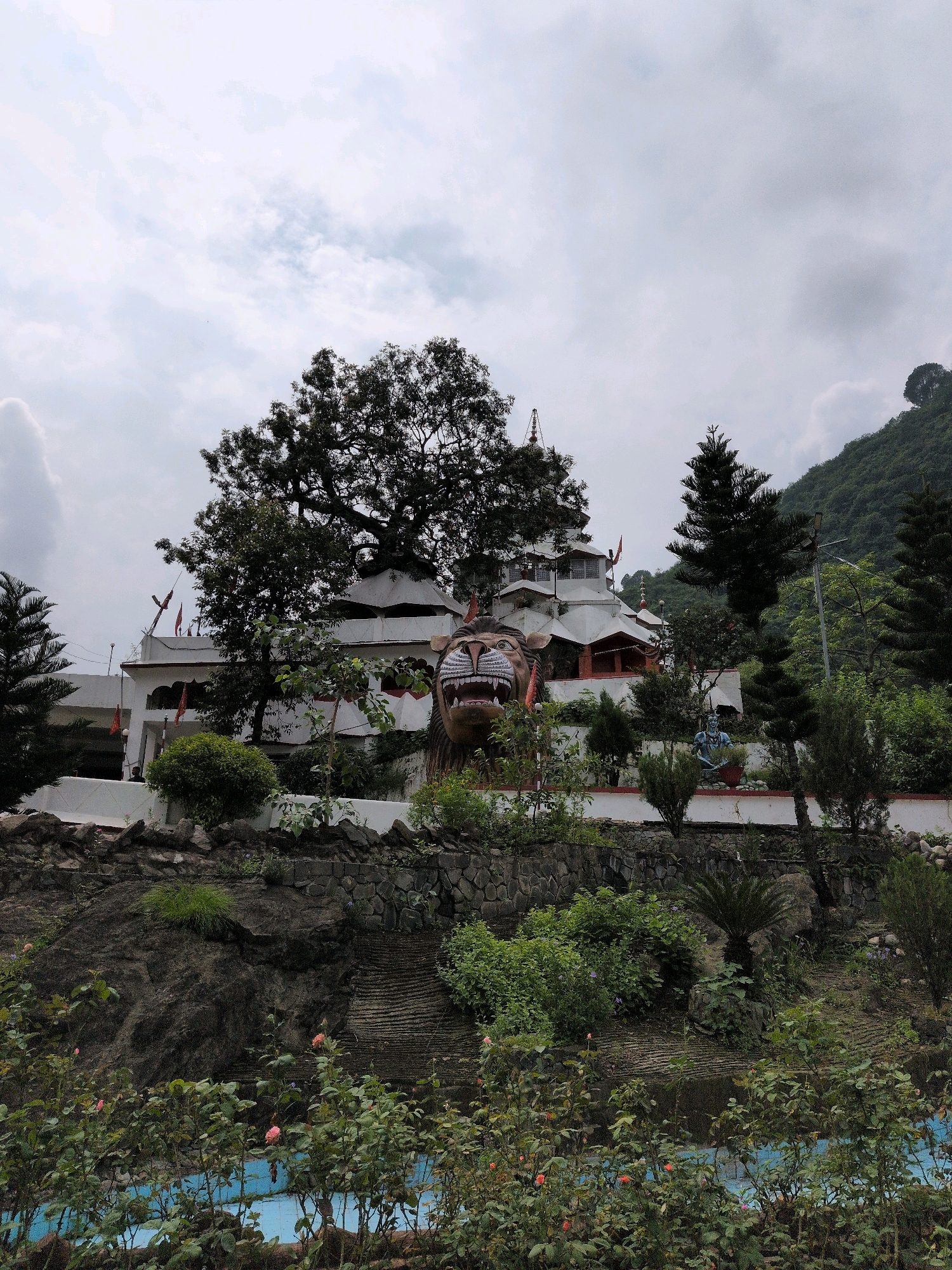 The image shows a religious building with white walls and a tiered roof structure situated on a hillside. In the foreground, there is a large, fierce-looking statue of a lion's head with its mouth open. The building is adorned with several flags, and there are trees and greenery surrounding it, as well as a statue of a seated figure, likely a deity, in the middle ground. A body of blue water, possibly a pool or pond, is visible in the lower foreground, surrounded by rose bushes. The scene is set against a backdrop of a cloudy sky and a lush green mountain slope on the right side of the image. This appears to be a Hindu temple located in Mandi, India, likely a place of worship and tourism.