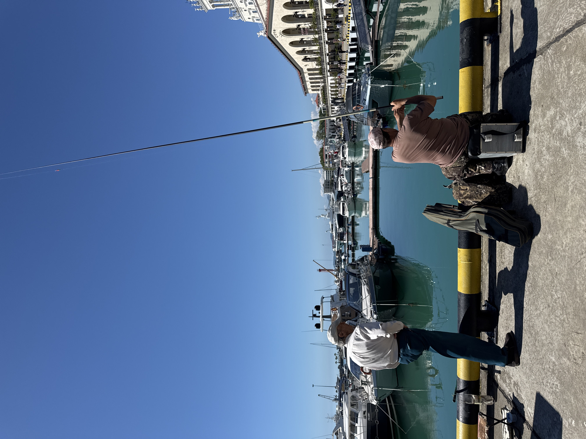 The image, correctly oriented, depicts a sunny day at a marina in Sochi, Russia, where two men are fishing from a concrete pier.
In the foreground, a man wearing a light-colored long-sleeved shirt and dark trousers stands with his back mostly to the camera, looking towards the water. A plastic water bottle rests on the pier near his feet. Further along the pier, a second man is seated on a dark box, actively holding a fishing rod extended over the calm, turquoise-green water. He wears a brown shirt, camouflage pants, and a light-colored cap. Additional fishing bags or cases are visible on the pier beside him.
The marina is bustling with numerous boats and yachts of various sizes docked along the pier and across the basin. The concrete pier itself features prominent yellow and black striped bollards. In the background, beyond the boats, stand several multi-story white buildings characterized by arched windows and balconies, suggesting a waterfront promenade or resort area.
The sky is a clear, deep blue with no clouds, indicating bright, sunny weather. The strong shadows cast by the men and the bollards suggest it is likely mid-day or early afternoon. On the hull of one of the boats docked in the marina, the registration number "P-04-001" can be faintly observed. The overall scene conveys a sense of peaceful leisure and outdoor activity.