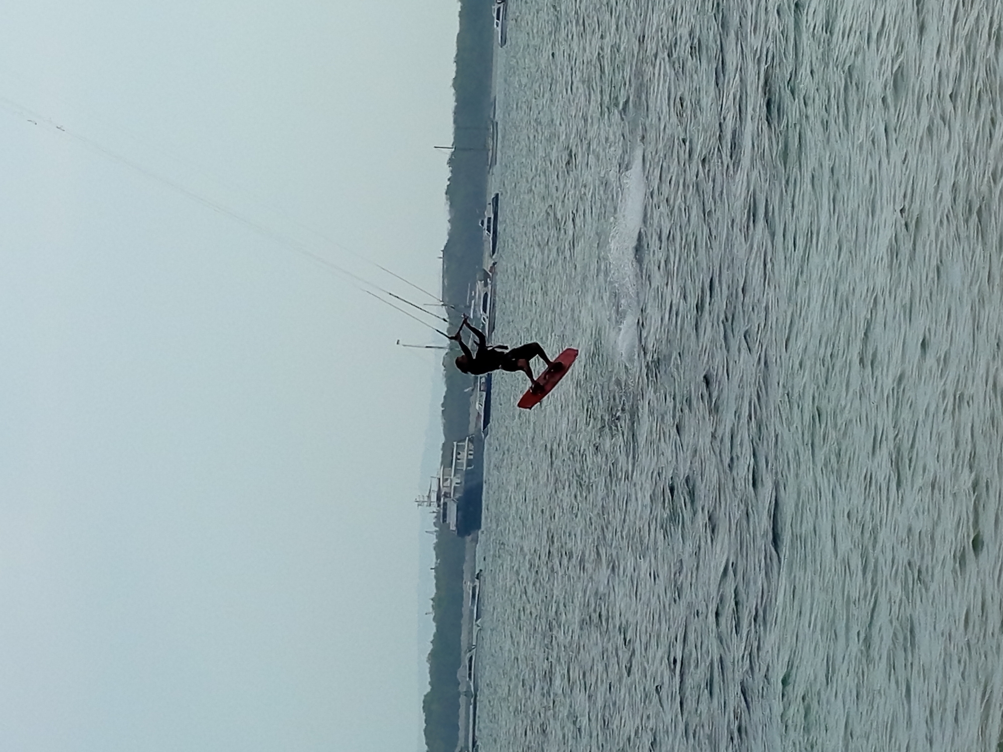 A silhouetted individual kiteboards on a body of water. The kiteboarder is in mid-air, performing a jump, with their kite connected via lines to their harness. The water has visible ripples and a small spray of water trails behind the board. In the background, several boats are anchored or moored, and a treeline with a distant hill forms the horizon. The sky is overcast and light blue. This scene is consistent with locations in Denpasar, Indonesia, such as the waters off Sanur Beach, known for its kiteboarding activities and boat traffic.