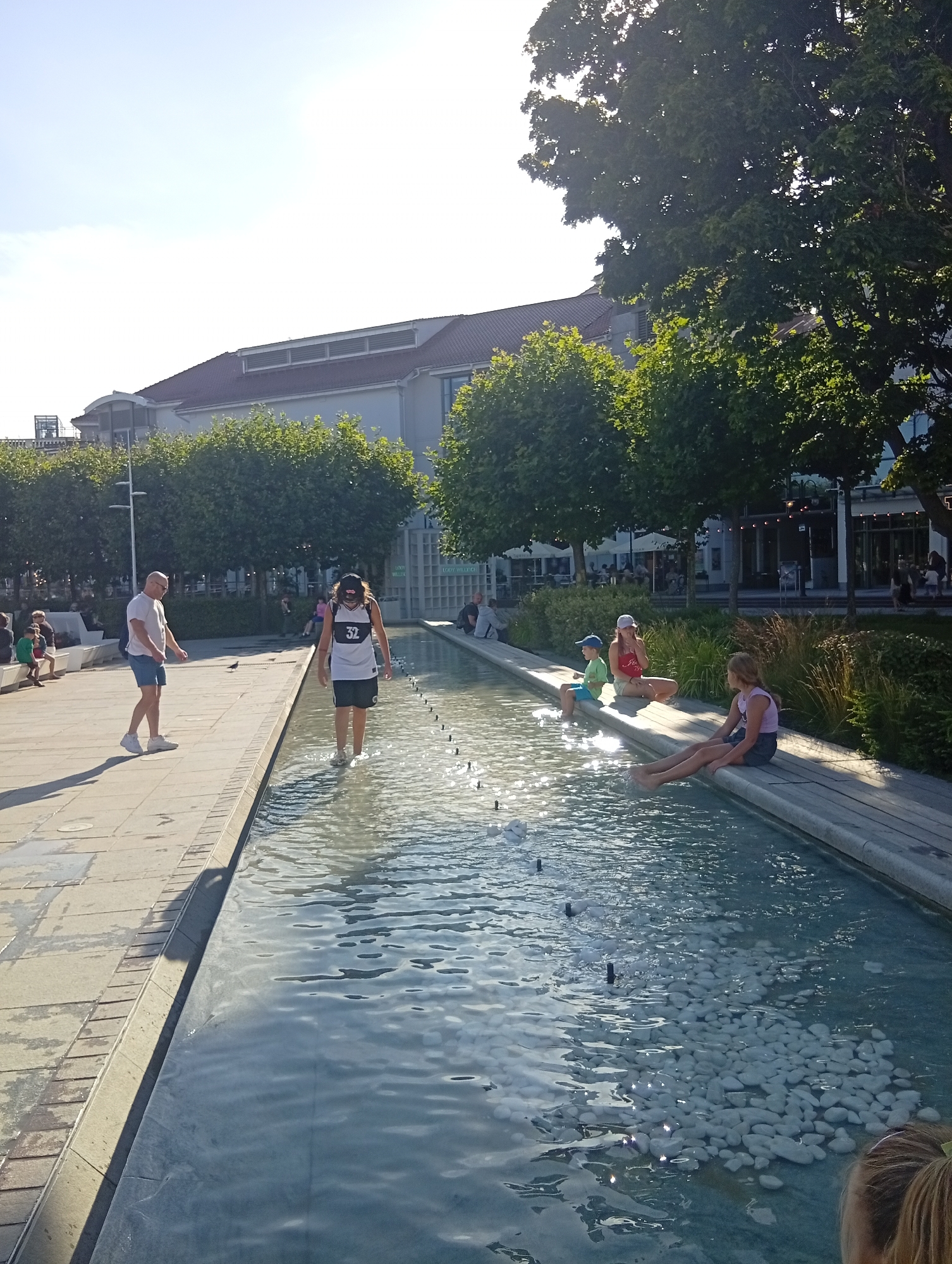 The image depicts a sunny outdoor scene, likely in the city of Sopot, Poland, featuring a shallow water feature with a paved walkway adjacent to it. A man in a white t-shirt and blue shorts stands on the walkway, facing a person in the water. In the water, a person wearing a white tank top with the number "32" and black shorts is walking through the shallow water. Further along the water feature, several people are seated on the edge. On the right side, a young girl in a pink tank top and denim shorts sits with her legs in the water, next to a boy in a green t-shirt and blue shorts. Another girl, wearing a light purple top and dark shorts, is also seated with her legs submerged. The background shows trees, a building with a red roof, and other individuals in the distance. The water feature is lined with white pebbles and has small jets of water.