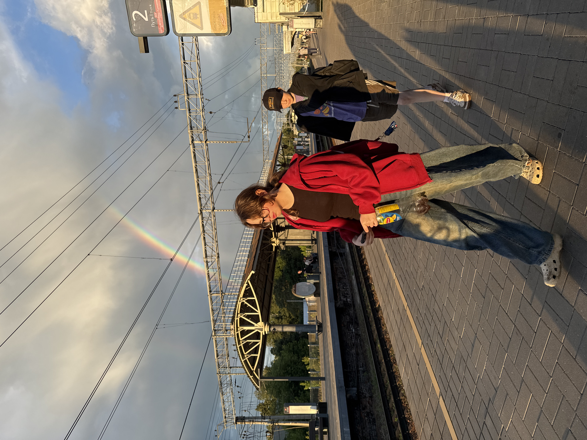 The image depicts a train platform in Bykovo, Russia, bathed in the warm glow of late afternoon sunlight. A faint rainbow arcs across a cloudy sky, partially obscured by overhead train wires. Two individuals are walking along the platform. In the foreground, a person wearing a red hooded jacket, a black t-shirt, and loose-fitting jeans is walking with their head down, holding a bottle of iced tea and a phone. They are wearing white Crocs. Behind them, another individual in a dark jacket, a purple t-shirt, and shorts walks with a backpack slung over their shoulder and is wearing a black baseball cap. The platform is paved with grey bricks, and train tracks run alongside it. A covered waiting area with supporting pillars is visible further down the platform. Signage indicates "Track 2" and offers safety warnings in Russian. Trees and buildings can be seen in the background.
