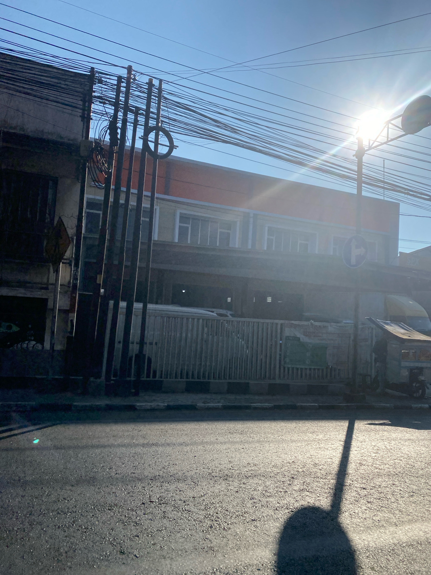 The image depicts a street scene bathed in bright sunlight, casting long shadows across the asphalt road. On the left, a weathered building with dark windows and a yellow diamond-shaped sign is partially visible. Adjacent to it are several dark, vertical poles supporting a dense network of overhead electrical wires. A circular object, possibly a coiled wire or part of a sign, hangs from one of the poles.
The background features a multi-story building with an orange upper facade and rectangular windows. In front of this building, a light-colored fence runs horizontally. A street sign with a blue circular background and a white arrow indicating a right turn is mounted on a pole in front of the fence. To the right, a partial view of a yellow vehicle and a cart with a thatched roof can be seen. The sky is clear and blue, with the sun prominently visible on the right, causing lens flare and intense brightness. The overall impression is an urban street under a strong, sunny sky, with a focus on infrastructure like utility poles and wires.