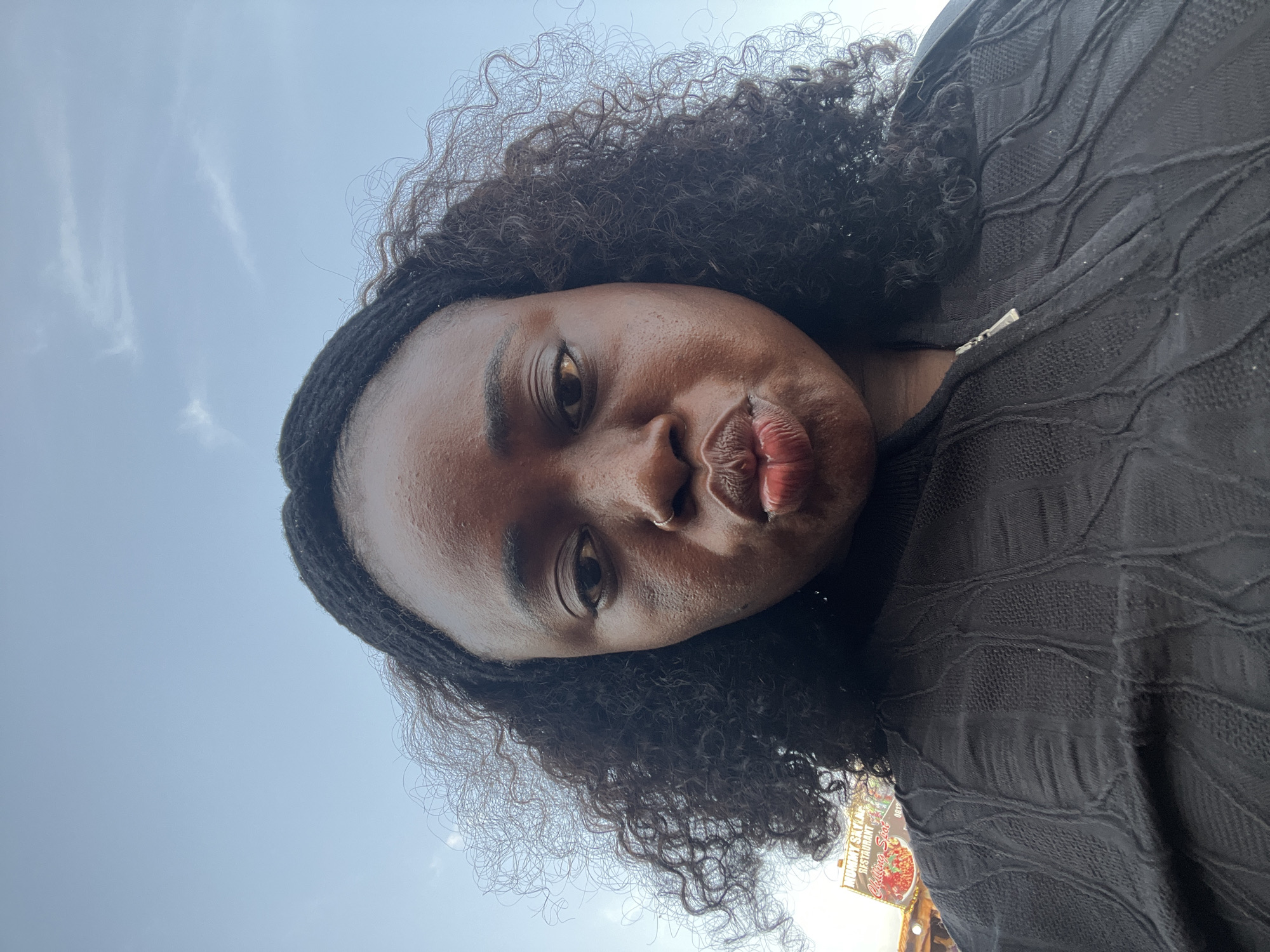 A close-up, low-angle selfie shows a Black woman with dark, curly hair and a black zip-up top. She is making a kissing face, with her lips pursed. Her skin has a light sheen. In the background, a sign for "MUMMY SKYLINE RESTAURANT" is partially visible, indicating the location is likely in Abuja, Nigeria. The sky is a pale blue with wispy clouds.