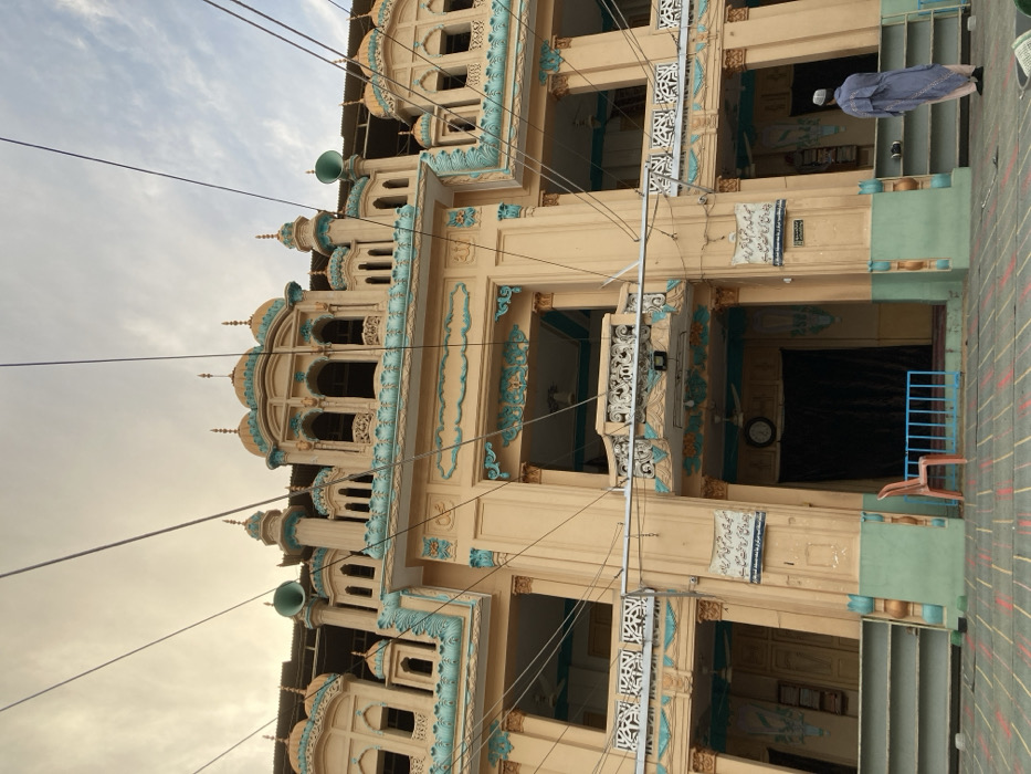 This image displays the ornate facade of a multi-story religious building, likely a mosque, situated in Quetta City Tehsil, Pakistan. The structure is primarily painted in a light yellow/cream hue, extensively decorated with turquoise and light blue trim, intricate carvings, and architectural details. Its upper sections feature numerous small golden-domed cupolas and minaret-like spires, arched window openings, and balconies with white patterned railings. Two green horn-shaped loudspeakers are affixed to the upper facade, and a corrugated metal roof is visible behind the highest domes.
Multiple dark electrical wires crisscross the sky and the building's facade. The sky overhead is overcast with brighter patches.
At ground level, the building exhibits three recessed sections, each with a dark interior. A single red plastic chair is positioned to the left of the central recessed area, which is fronted by a low blue metal barrier. To the left of this central section, a wall-mounted clock is visible above a dark curtained doorway. A white shelving unit is attached to the wall to the right of the central section. White rectangular signs with Urdu script are present on the facade, notably above the central area.
In the lower right foreground, a single male individual stands facing the building with his back to the camera. He is dressed in a long, light blue/grey robe and a white skullcap, positioned on patterned grey and black paving stones that constitute the ground surface. His posture suggests contemplation or preparation for a religious act.