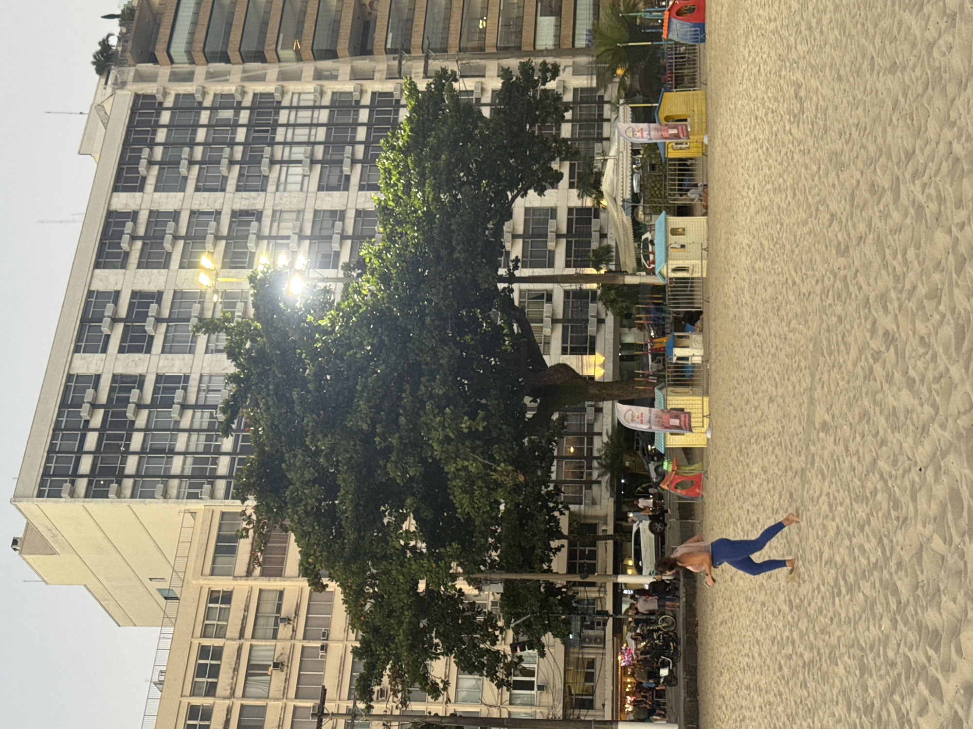 The image depicts a beach scene in Niterói, Brazil, with a woman jogging on the sand in the foreground. Behind her, a large tree stands in front of several multi-story buildings lining the street that runs along the beach. There are small, colorful playhouses set up on the sand, some with flags. A metal fence separates the play area from a sidewalk with people, motorcycles, and cars. The buildings are modern and appear to be apartment or hotel complexes. The sky is overcast.