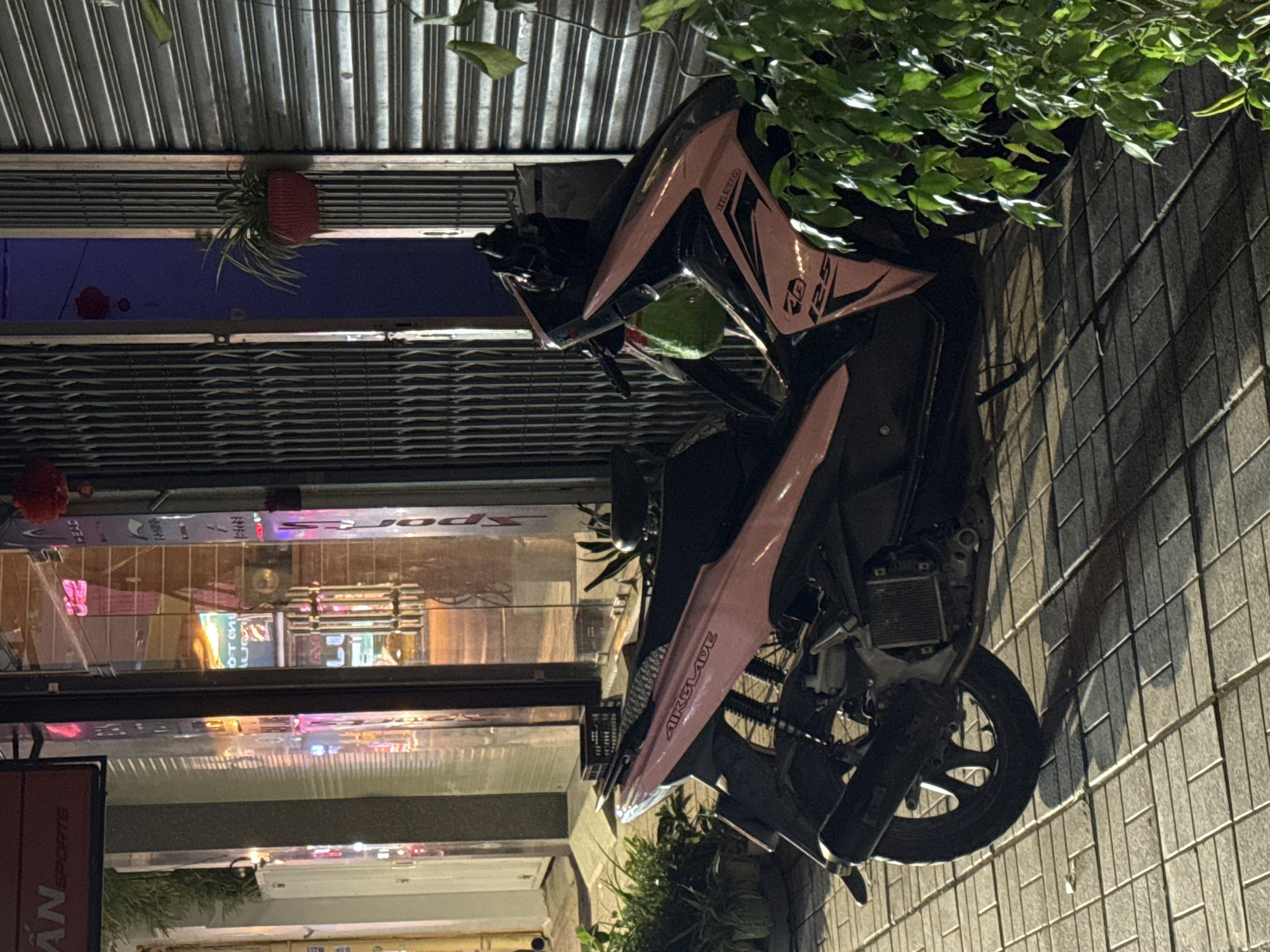 A pink and black Honda Airblade scooter is parked next to a building with a corrugated metal facade and a sidewalk. The scooter is the main subject, with its front fairing prominently displaying the "HONDA" and "AIRBLADE 125" logos. The scooter's color scheme is primarily pink with black accents.
The scene appears to be an urban setting in Nha Trang Ward, Vietnam, indicated by the architectural style and the presence of storefronts. The building has large glass windows that reflect the lights from within, suggesting it is a shop or commercial establishment. Some of the reflected text from the shop windows is visible, including what appears to be "SPORTS" and other Vietnamese characters.
A potted plant hangs from the metal facade above the scooter, adding a touch of greenery to the scene. Lush green leaves from an overhanging plant cascade down from the top right of the image, partially obscuring the scooter and the building. The lighting suggests it is evening or night, as the storefront lights are illuminated and cast reflections. The ground is a tiled sidewalk, with a shadow from the scooter cast upon it. The overall mood is that of a typical street scene in a bustling Vietnamese city.