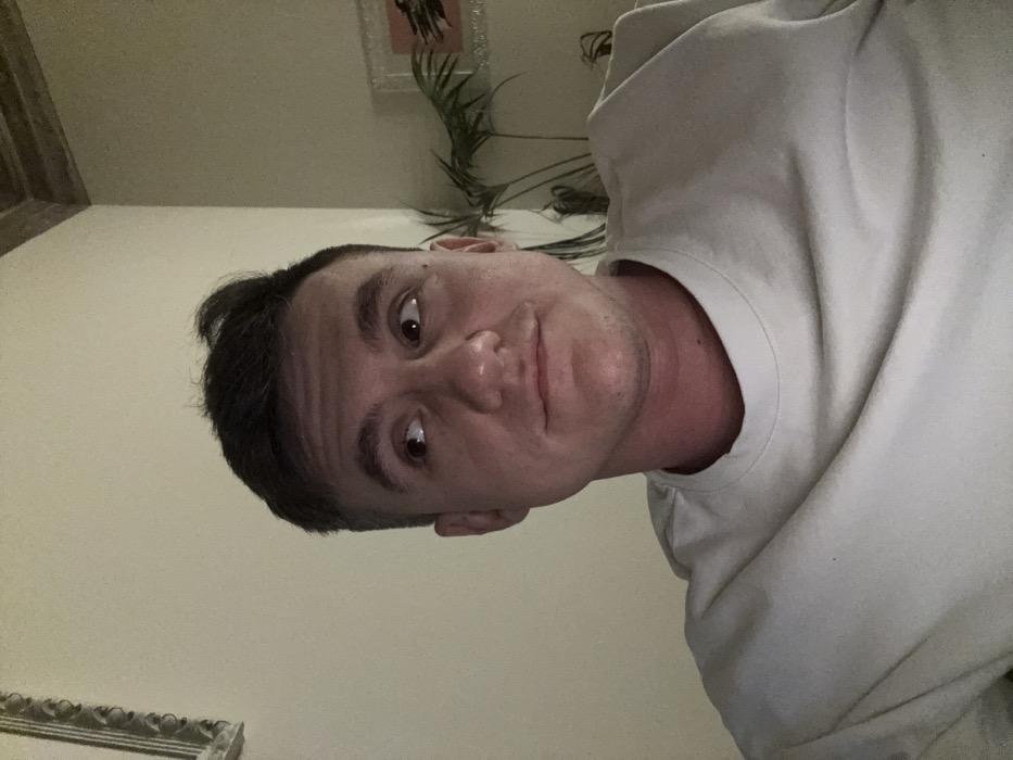 An adult male is captured in a head-and-shoulders portrait, positioned indoors in a room in Venice, Italy. He has short, dark hair and a light complexion, with some redness visible on his lower neck and upper chest. He is wearing a light-colored, possibly off-white or light beige, crew-neck shirt. His gaze is directed upwards and towards the viewer's left, with his lips slightly pursed in a subtle, neutral expression. The background consists of a plain, light beige wall. To the upper left, a portion of an ornate, possibly silver-toned, framed object is visible. In the upper right quadrant of the image, a section of a textured or paneled ceiling, along with a green leafy houseplant and a framed artwork featuring a prominent orange border or mat, are discernible. The scene is illuminated by warm, artificial light. No other individuals or interactions are visible.