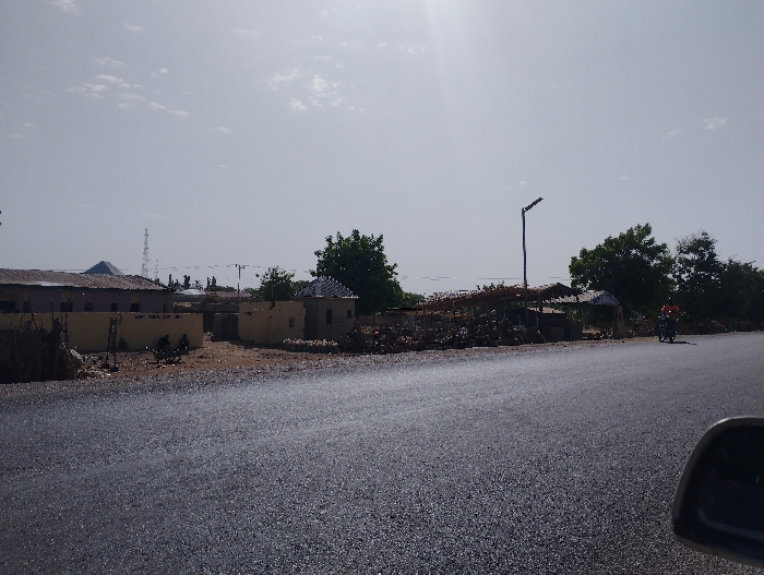 The image shows a paved road, with a single motorcycle driver visible on the right side of the road. There are buildings to the left, including a single-story building with a visible yellow wall, and other low-lying structures, some with corrugated metal roofs. There is a streetlight on the right side of the road, as well as trees behind the structures.  The sky is bright and slightly cloudy.  The image was taken in Kumo, Nigeria, at coordinates (10.047, 11.202).  The image is taken at a slight angle to the road, from the passenger seat of a vehicle looking out.
