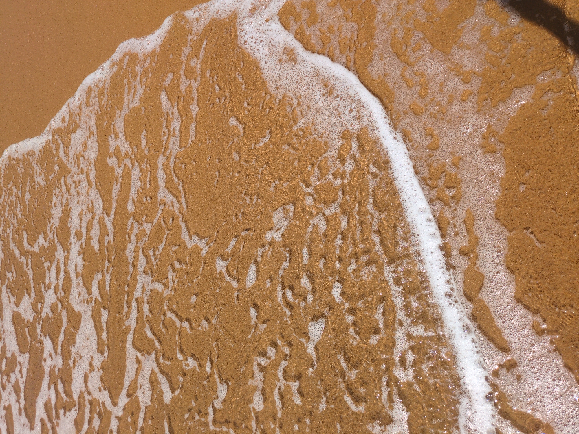 The image depicts a close-up view of the shoreline at a beach. Golden sand is visible, partially submerged by foamy seawater. The water's edge is marked by a line of white foam, creating a transition between the wet sand and the dry sand further inland. The scene is likely located in a coastal area, potentially in South Quy Nhon Ward, Vietnam, given the prompt's indication of the location. No people or other specific objects are visible, the primary subjects are the sand and the water.