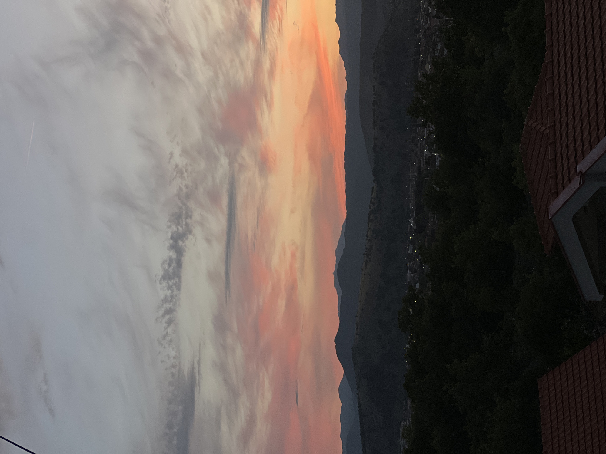 The image captures a twilight scene over a city, likely Podgorica, Montenegro, given the provided context. The sky is painted with soft hues of pink and orange from a setting or rising sun, with wispy clouds stretching across the horizon. Below the vibrant sky, a series of dark, undulating mountains form a silhouette against the colorful sky. In the mid-ground, the city lights begin to twinkle, interspersed with dense, dark foliage that obscures much of the urban landscape. In the foreground, the tiled rooftops of residential buildings are visible, with one roof on the left in shadow and another partially illuminated on the right. There are no people or specific actions depicted in the image, focusing entirely on the natural beauty of the sky and the subdued urban environment.