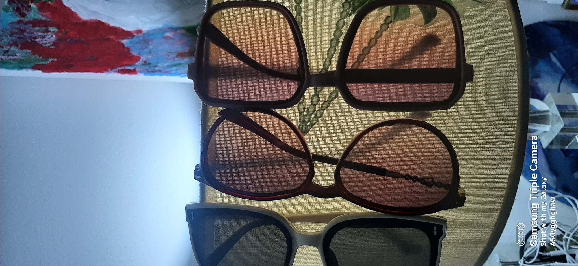The image depicts a still life composition featuring three pairs of sunglasses arranged on a textured, woven surface, possibly a lampshade. The sunglasses vary in frame color and lens tint. The lighting suggests an indoor setting, with a bright light source emanating from behind the objects, creating shadows and highlighting the mesh-like texture of the lenses. A portion of a painting with abstract red, blue, and green elements is visible in the upper right background, and a glass table lamp base is present in the lower right. A watermark at the bottom left indicates the image was taken with a "Samsung Triple Camera Shot with my Galaxy A50yyghghaw." The scene is not identifiable as being in Dubai, United Arab Emirates, or any specific city.