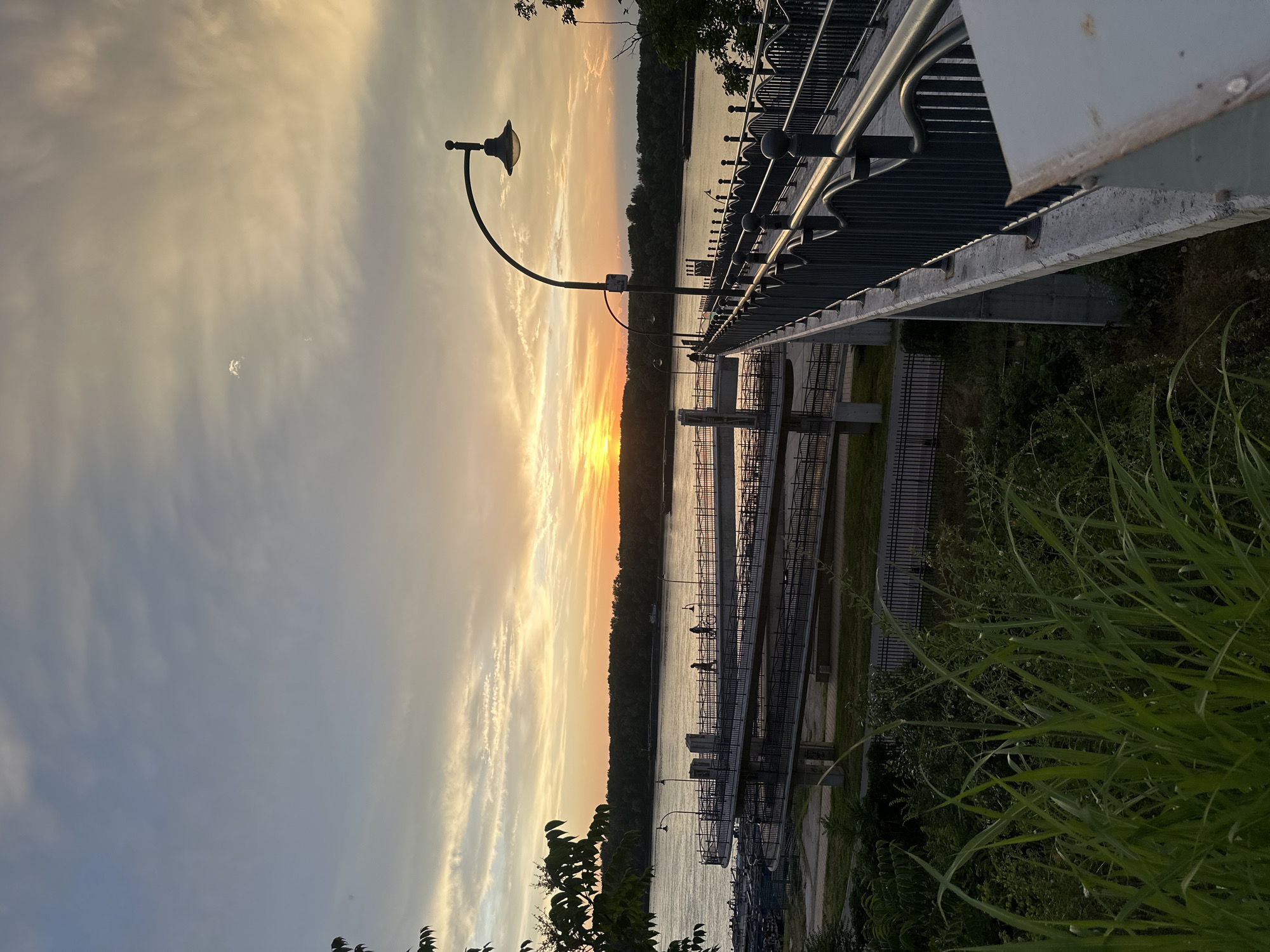 This is a vertical, eye-level shot of a bridge during sunset in Ruse, Bulgaria. The sky is filled with a gradient of colors, from soft blues at the top to warm oranges and yellows near the horizon. Clouds are scattered across the sky, catching the light of the setting sun. The bridge itself is made of concrete and has a dark metal railing with decorative elements. A lamppost with an antique-style fixture arches over the railing. Several people can be seen walking and cycling on the bridge, and a barge is visible on the river in the background. The foreground is dominated by lush green grass and foliage. The river, likely the Danube, is wide and calm, reflecting the colors of the sky. The overall atmosphere is peaceful and serene.