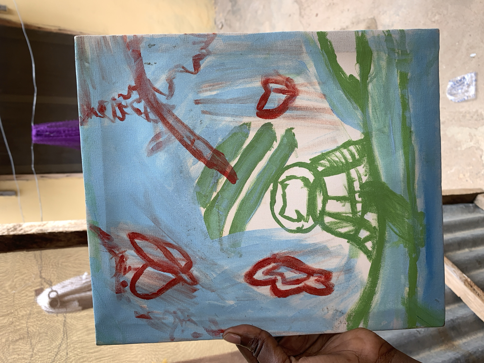 A dark-skinned hand holds a small canvas painting outdoors. The painting is an abstract piece, featuring a light blue background with streaks of white. It contains various shapes and lines in red and green paint. Red shapes resembling hearts or abstract forms are scattered across the canvas, while green shapes include linear strokes and a cage-like form. The background outside the canvas is blurry, but shows a window with purple material, a wooden frame, and a corrugated metal surface. The location is Rafin Par, Nigeria.