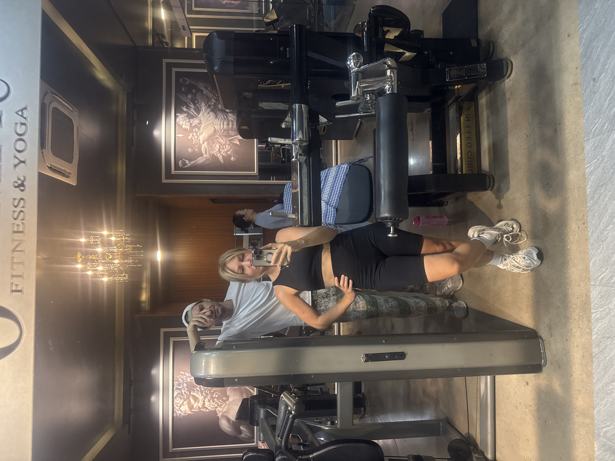 The image shows a gym located in Nha Trang Ward, Vietnam, with a large mirror reflecting the scene. In the foreground, a woman is taking a selfie, wearing a black sports bra and black biker shorts, with white sneakers and white socks. To her left and slightly behind her, a man is leaning against a piece of gym equipment, wearing a white t-shirt, a white baseball cap, and patterned pants. The gym appears to be well-equipped with various machines and has decorative elements such as framed artwork on the walls and a chandelier on the ceiling. Another woman is visible in the background, sitting and looking away from the camera. The overall lighting suggests an indoor environment with overhead illumination.