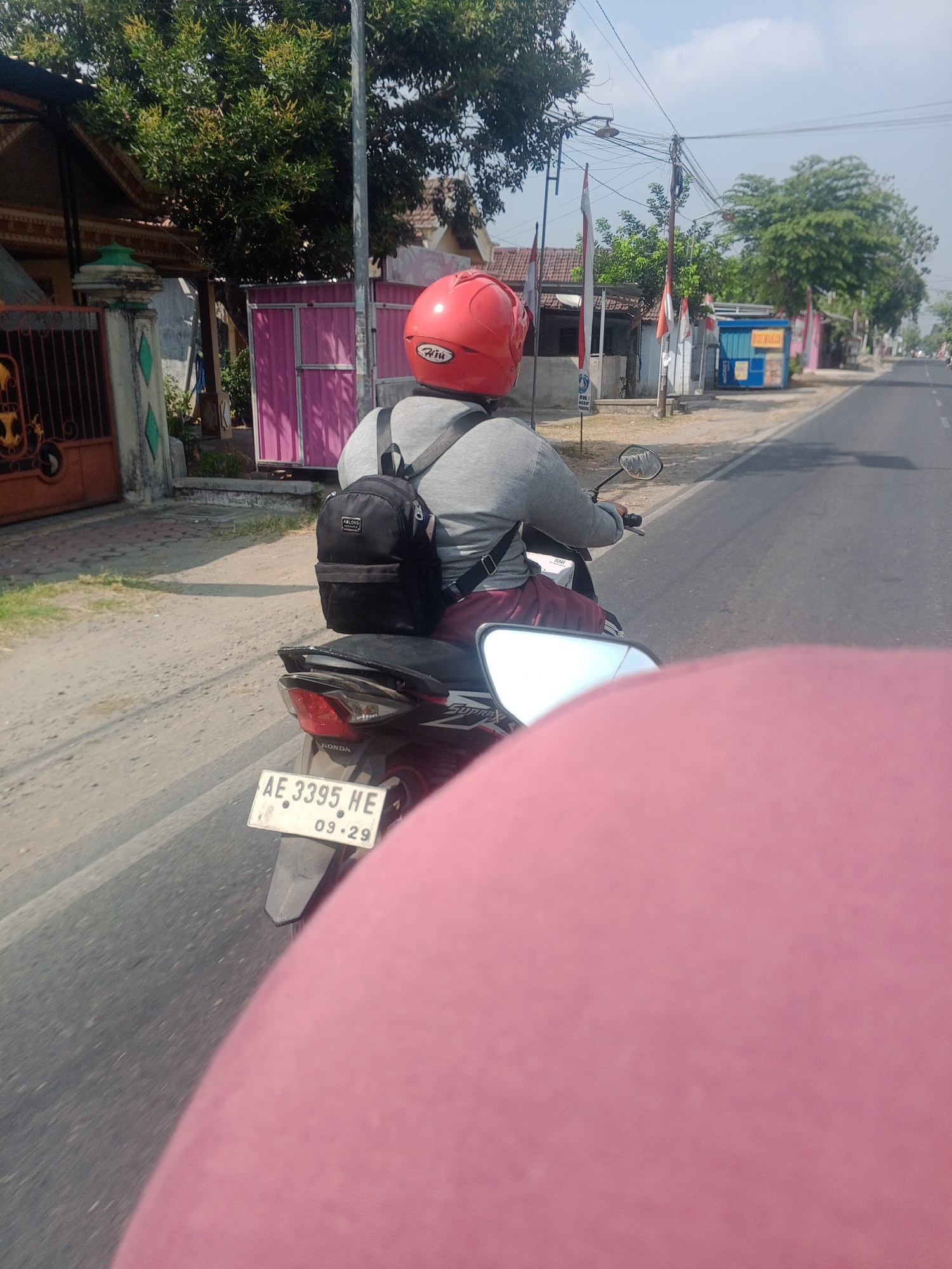 In Plalangan, Indonesia, a person wearing a red helmet and a gray long-sleeved shirt rides a motorcycle down a paved road. The rider has a black backpack on their back. The motorcycle has a license plate that reads "AE 3395 HE 09.29". The scene is set during the day under a clear sky. The road is bordered by a dirt shoulder and houses with trees and fences. A pink object, likely another person or clothing, is visible in the foreground, obscuring the lower part of the image.