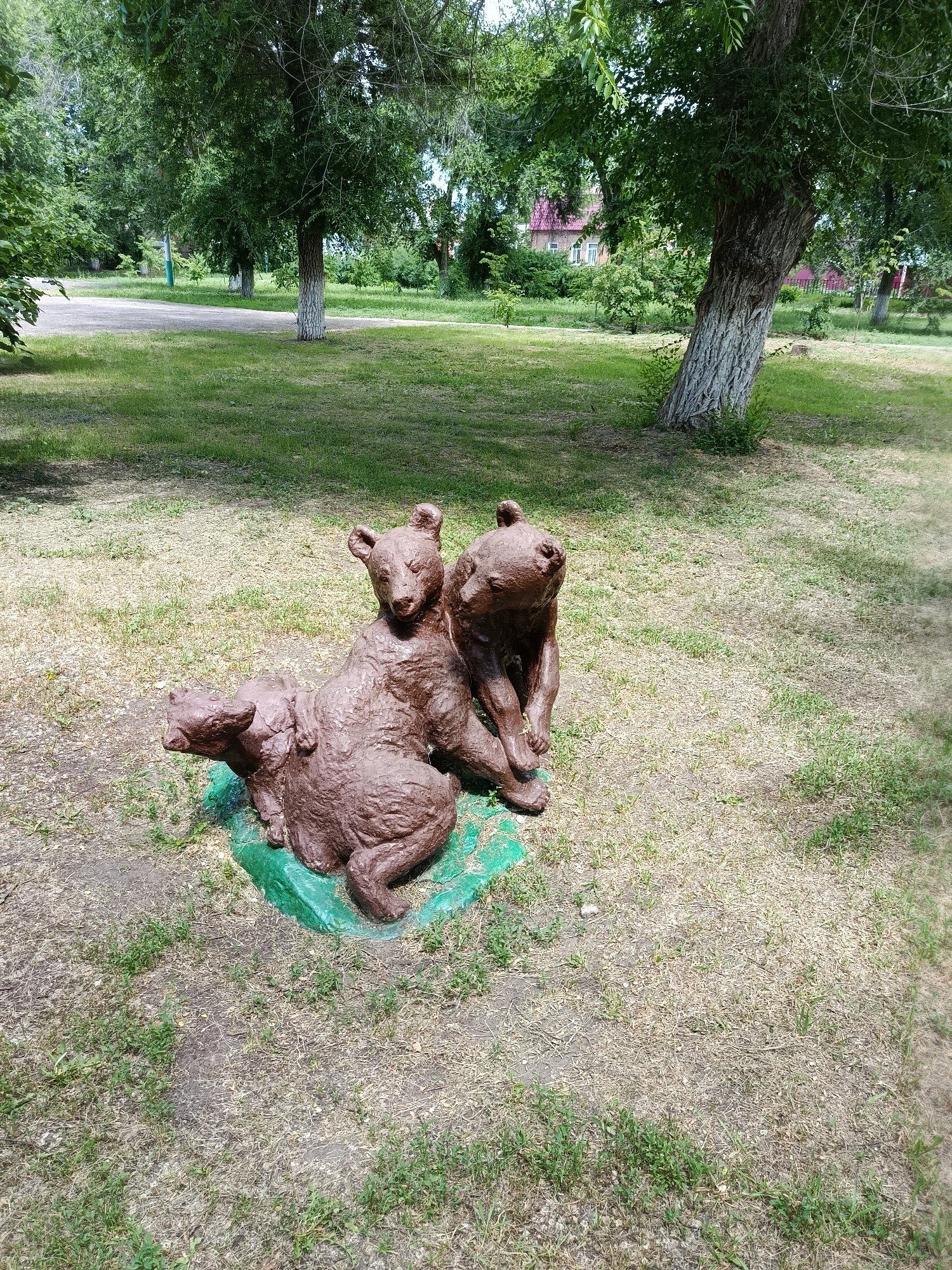 A sculpture of three bear cubs is situated on a patch of grass in a park in the city of Marx, Russia. The cubs are depicted huddled together, with one cub appearing to be sitting on another. They are made of a reddish-brown material and are placed on a green, irregularly shaped base. The park setting features mature trees with green foliage and a paved pathway in the background. The ground around the sculpture is covered with a mixture of dry grass, dirt, and scattered green weeds. In the distance, buildings with red roofs and fences are visible among the trees. The lighting suggests it is daytime with some dappled sunlight filtering through the leaves.