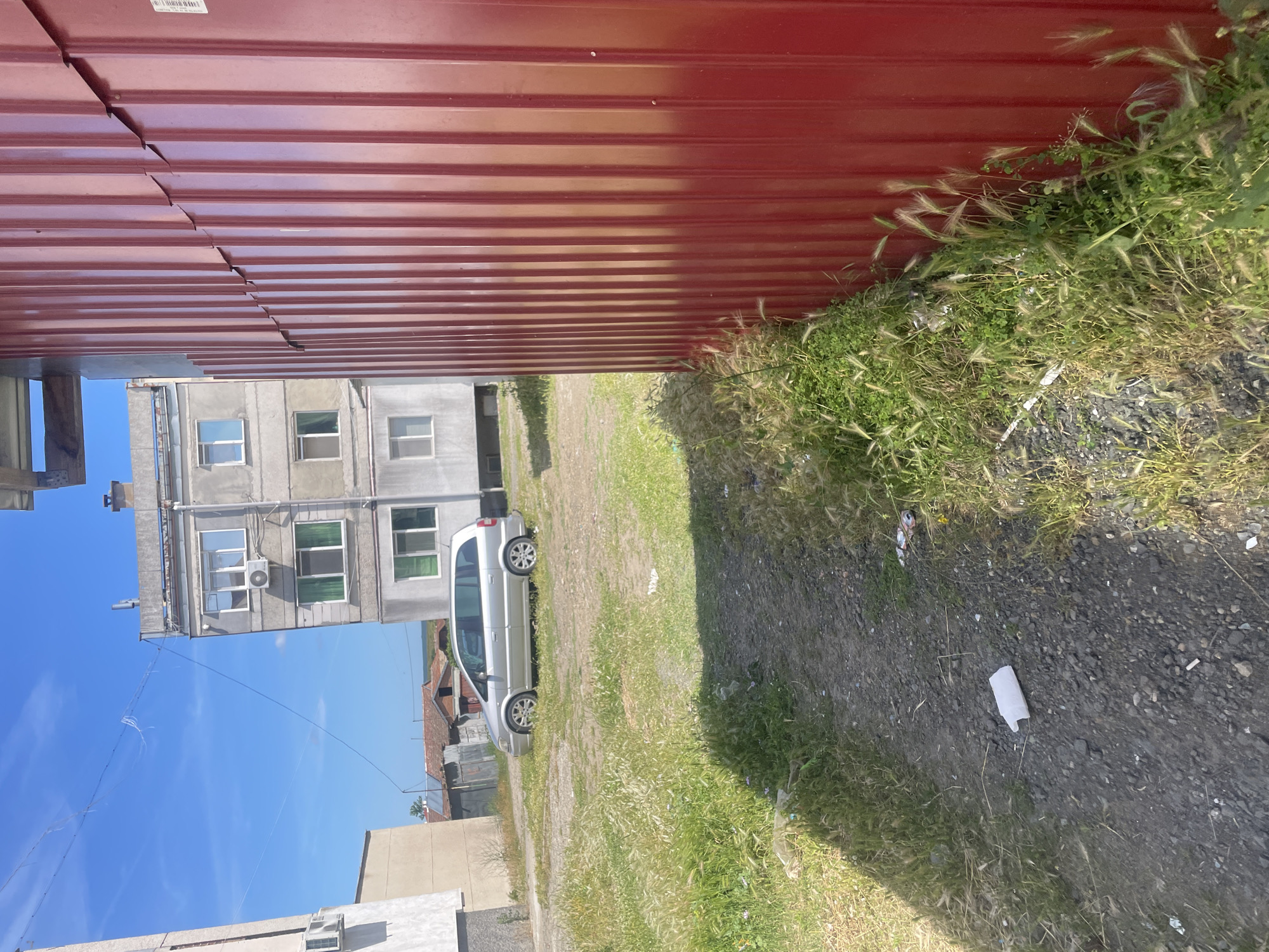 In Debelt, Bulgaria, a silver minivan is parked on a dirt lot next to a multi-story apartment building. The building has a gray facade and several windows. A red corrugated metal fence runs along the right side of the frame, partially obscuring the apartment building and a portion of the sky, which is blue with some wispy clouds. The ground is covered in dirt, gravel, and patches of green grass and weeds. There is some scattered litter on the ground, including a white plastic object and a crushed can. The scene appears to be an outdoor urban environment.