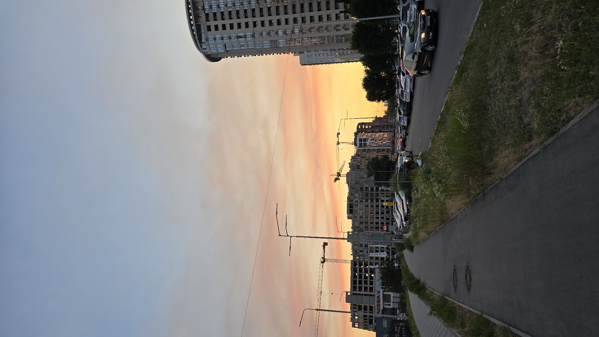 The image depicts an urban scene at sunset, likely in Kyiv, Ukraine. The sky is ablaze with vibrant hues of orange, pink, and yellow, indicating a dramatic sunset. The foreground features a paved path, likely a sidewalk or bicycle lane, bordered by a grassy verge and then a road where numerous cars are parked. Beyond the road, a cityscape unfolds with several multi-story buildings, some of which appear to be under construction, indicated by the presence of cranes. One building has a prominent "NISSAN" sign. The overall atmosphere is one of a developing urban area as the day draws to a close. The lighting suggests it is late afternoon or early evening.