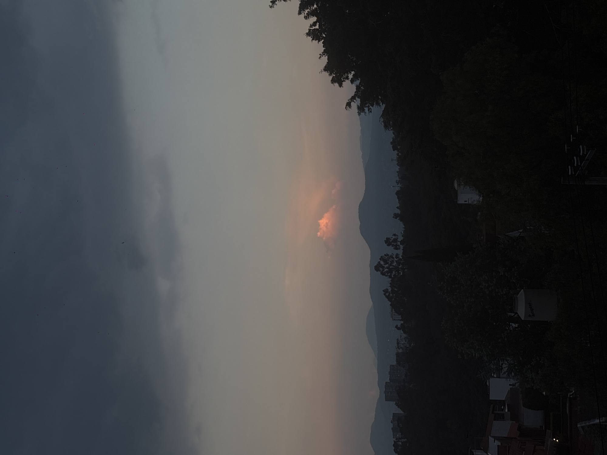 The image shows a dusky sky over what appears to be Mexico City, Mexico. A line of hills or low mountains can be seen in the distance. The right side of the image shows trees, rooftops of buildings, and electrical wires, indicating a residential or urban area. The left side of the image depicts a dark, cloudy sky. The light in the sky is muted and suggests a sunset or sunrise.