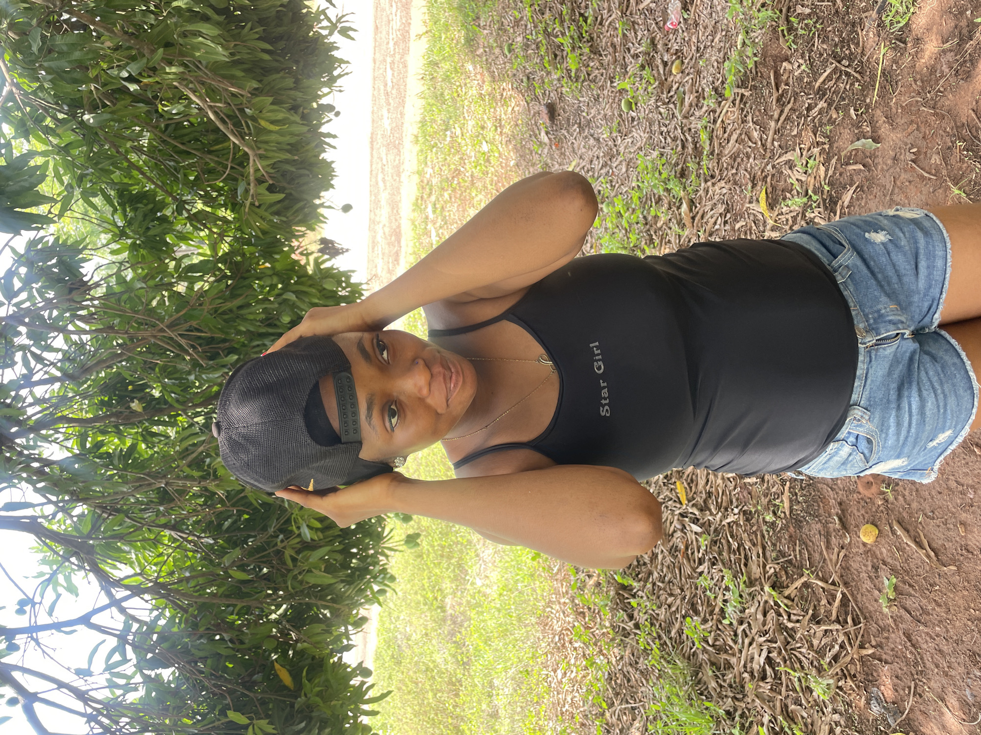An eye-level shot depicts a woman outdoors in Ogbe, Nigeria. She is centered, wearing a black tank top with "Star Girl" printed on it, distressed denim shorts, and a dark mesh baseball cap. Her hands are positioned near the hat. The background consists of green foliage, likely from trees and ground vegetation, and a patch of lighter ground in the distance. The ground near the woman is covered in dry leaves and some green plants.