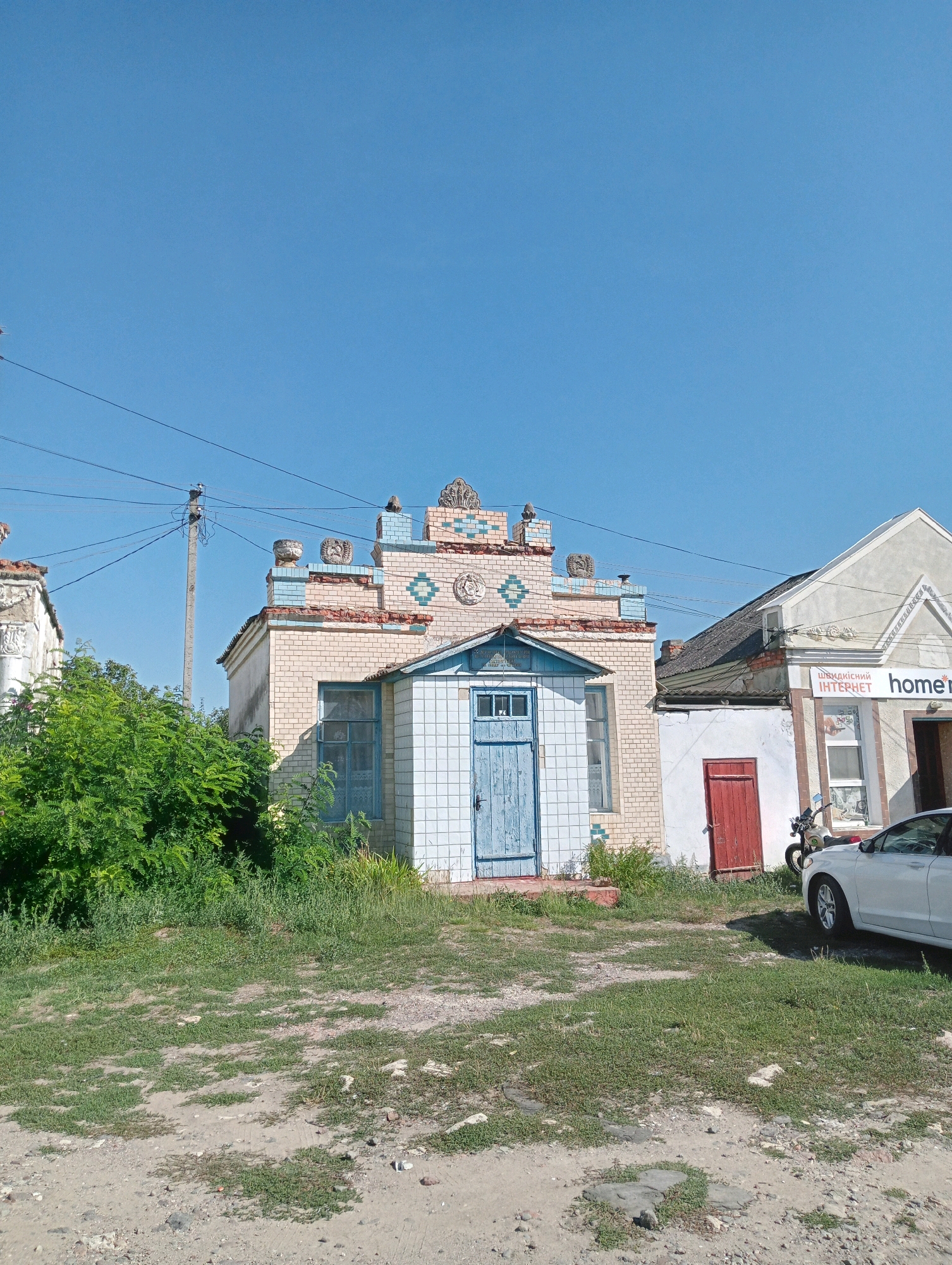 In Bronnytsia, Ukraine, on a bright, clear day, a light beige brick building with decorative blue and white tile accents stands prominently. The building features a light blue door with a window above it, and a single window with blue panes on its left side. To the right of this structure, a second building with a lighter colored facade is visible, displaying an advertisement for "ШВИДКІСНИЙ ІНТЕРНЕТ" (High-Speed Internet) and the word "home". A white car is parked in front of this second building, and a motorcycle is also visible nearby. Lush green bushes and overgrown grass surround the buildings, with a dirt and gravel ground surface. The sky above is a clear, vibrant blue, with power lines and a utility pole present in the upper left portion of the image.
