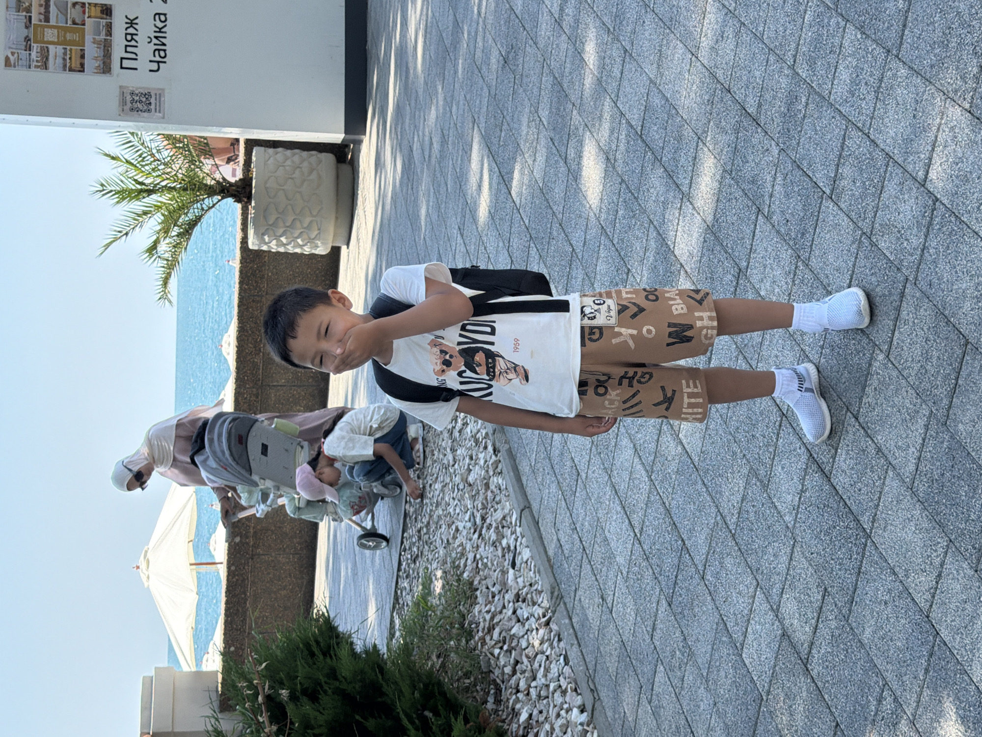 The image shows a young boy standing on a paved walkway, covering his mouth with his hand, as if suppressing a laugh or sneeze. He is wearing a white t-shirt with a graphic print of bears and text, and brown shorts with similar lettering. He also has a black backpack on and is wearing white sneakers and white socks. In the background, a woman in a hijab pushes a stroller with a child inside. The scene appears to be outdoors, near a body of water, likely the Black Sea, given the palm tree and beach umbrellas visible. The text on a white sign in the background, "Пляж Чайка 2," indicates the location is Chaika Beach 2 in Sochi, Russia. The walkway is made of grey rectangular pavers.