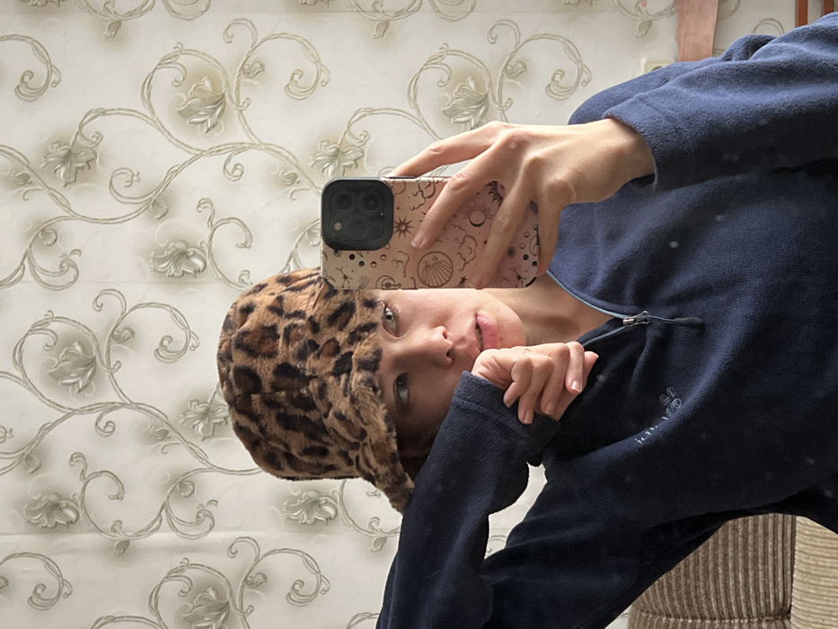 A woman is taking a selfie in a mirror. She is wearing a leopard print hat and a dark blue fleece jacket. Her phone, with a patterned case, is held in front of her face. The background features a patterned wallpaper in light shades and a hint of a wooden structure. The overall composition is slightly tilted.