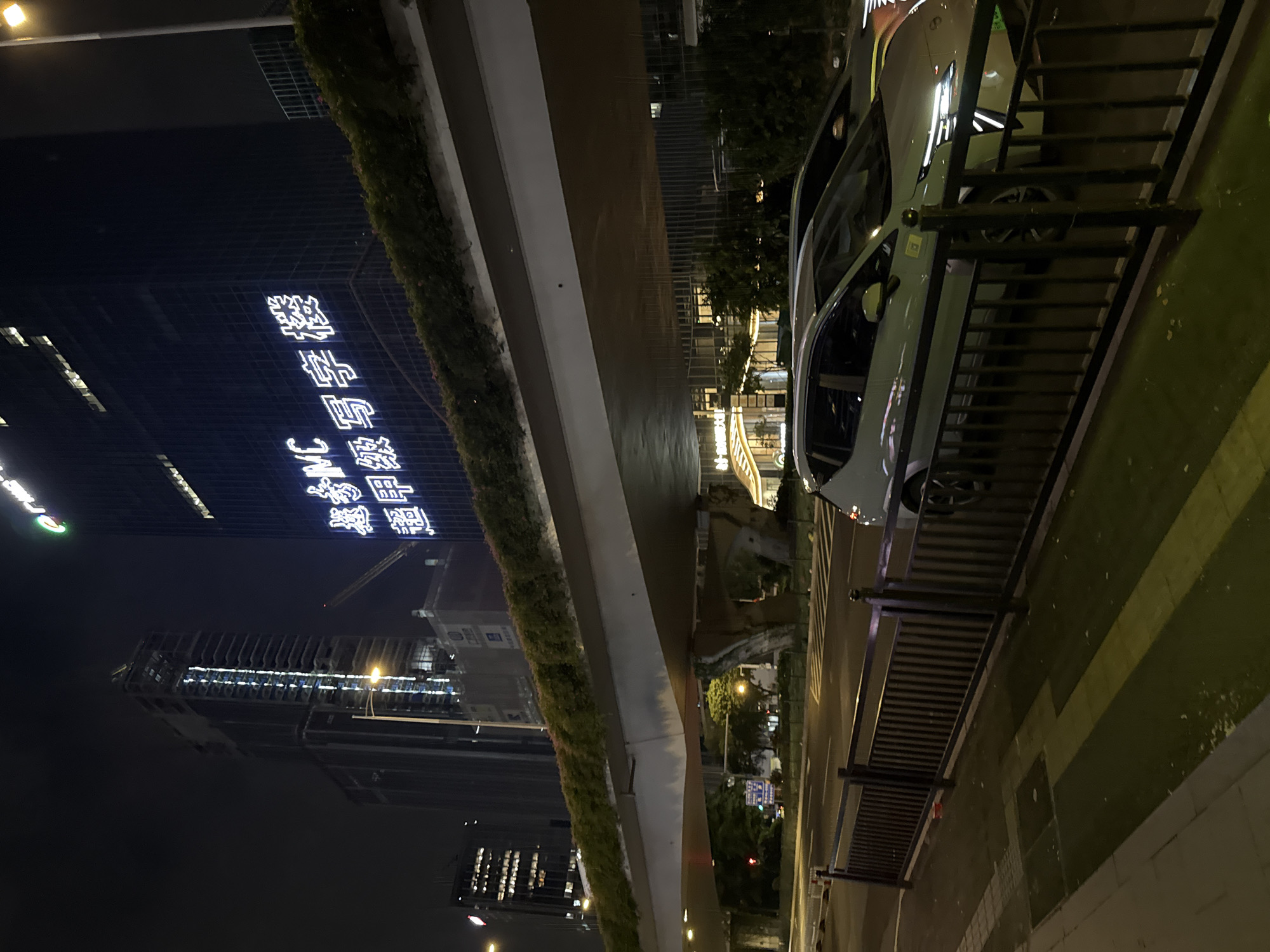 The image captures a nighttime urban scene in Huangpu District, China, dominated by a modern overpass casting shadows on a street below. Tall buildings rise in the background, with one illuminated by bright white neon signs displaying Chinese characters and the letters "IMC." The overpass itself is lined with lush green foliage. A light-colored car is parked on the street next to a black metal fence. The street is lit by streetlights and the glow from the buildings. The overall impression is one of a bustling, contemporary city environment after dark.