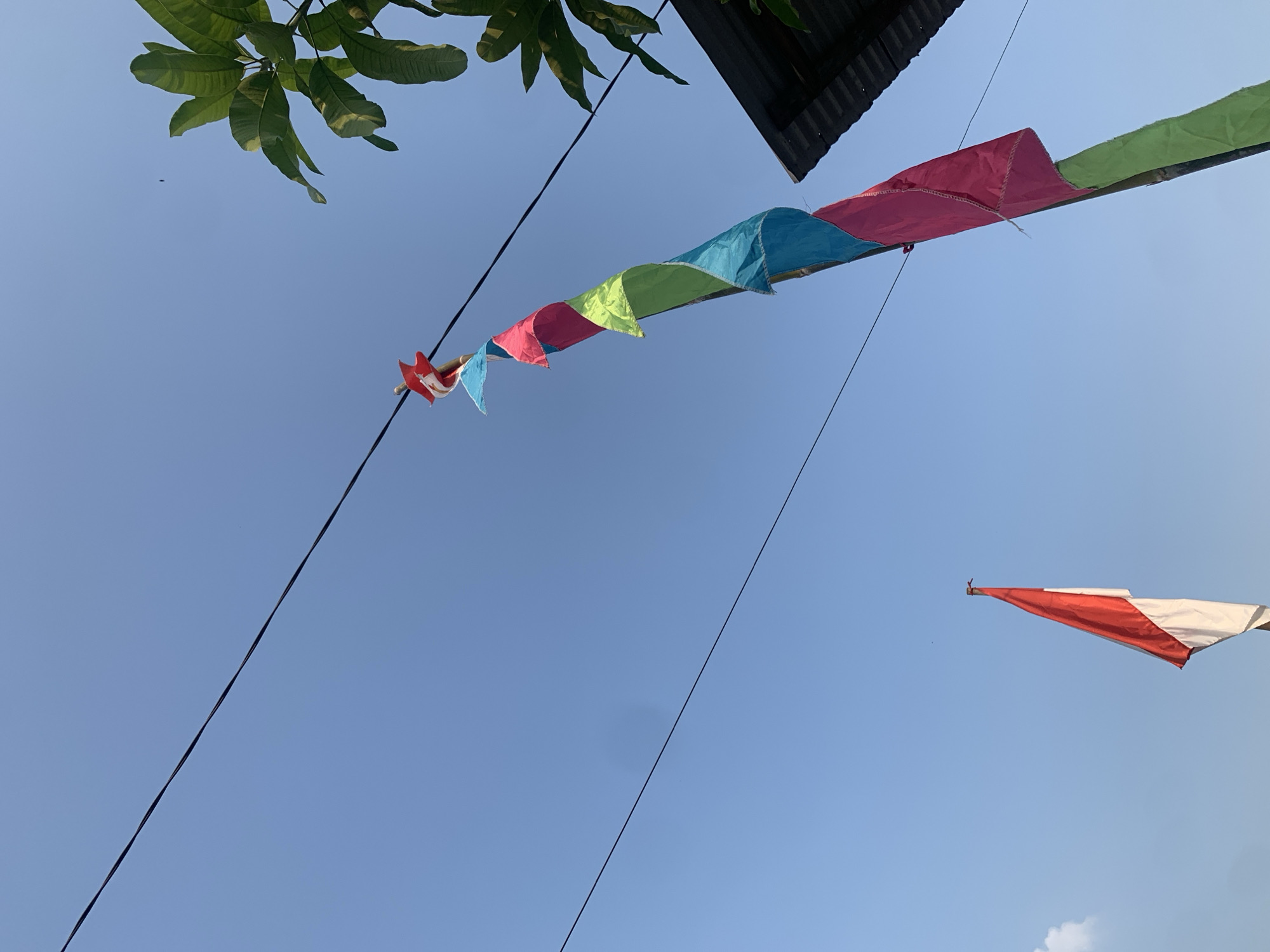 The image shows a clear blue sky above the city of Palembang, Indonesia, with two flags and power lines visible. On the left, a flag with red and white horizontal stripes, likely the Indonesian flag, stands tall. To the right, a tall bamboo pole is adorned with a string of colorful triangular flags in shades of pink, blue, and green, topped with a red star-shaped ornament. Green foliage from a tree extends from the upper right corner, and a dark corrugated roof is partially visible in the lower right. Power lines crisscross the sky, with one running diagonally from the top left to the bottom right. The scene suggests an outdoor setting, possibly a residential area or a public space decorated for a celebration or event.