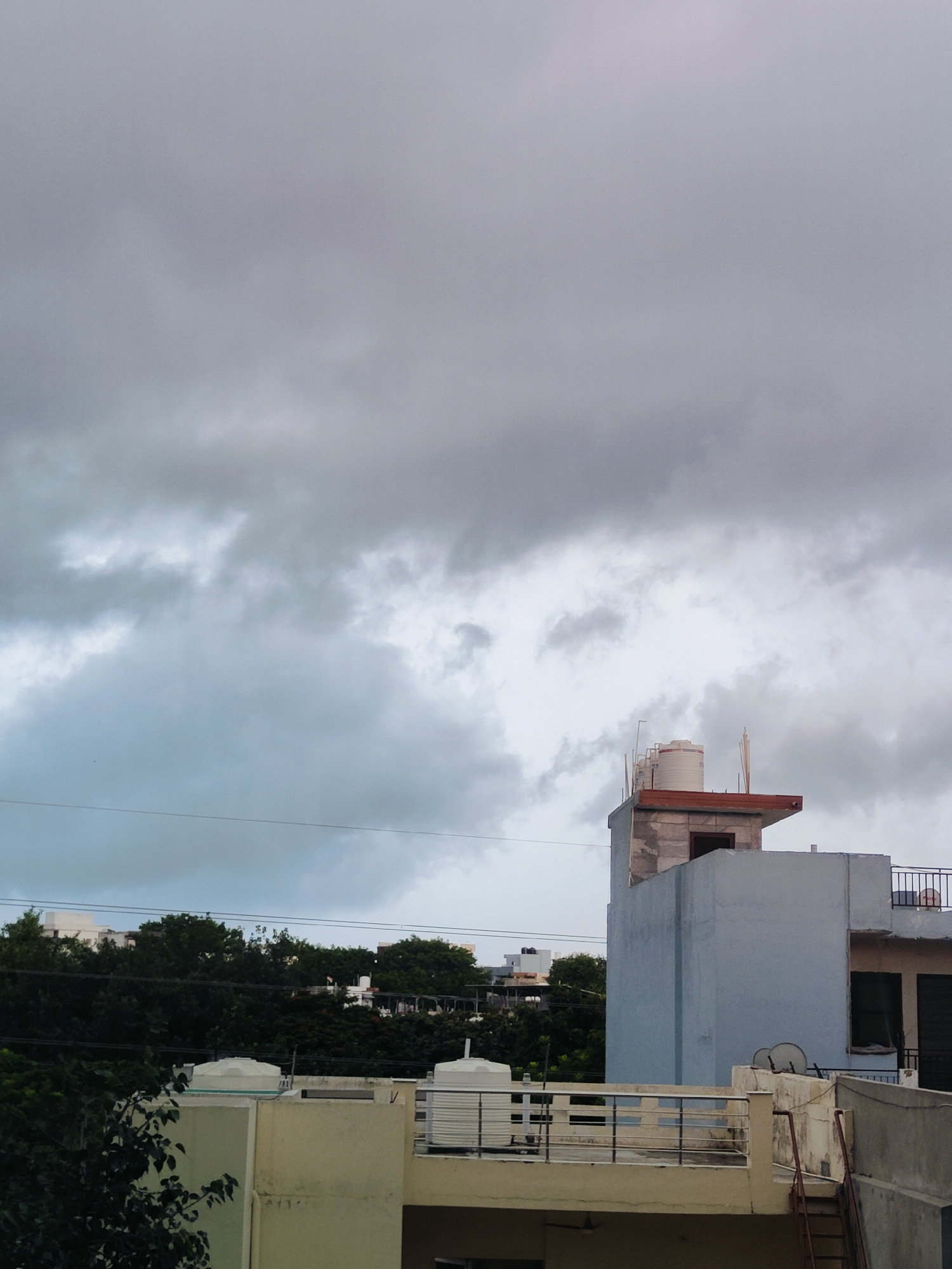 The image depicts a scene from Gurgaon, India, on a cloudy day. The foreground features the rooftops of several buildings. A pale yellow building with a light blue trim occupies the lower left portion of the frame, with a white railing and a black metal fence on its roof. Next to it, a light gray concrete structure rises, topped with a white circular water tank and a railing. Further to the right, a taller, light blue building is visible, with a dark recessed window and a section of a dark gray wall. A small satellite dish is mounted on the side of the blue building. A rusty red ladder leans against a concrete structure in the lower right corner. In the mid-ground, a dense line of dark green trees separates the foreground buildings from a row of white structures in the distance. The sky above is filled with heavy, grey clouds, suggesting an overcast or impending rainy day. A thin black wire stretches horizontally across the mid-section of the image, possibly a utility line. No people are visible in the frame.