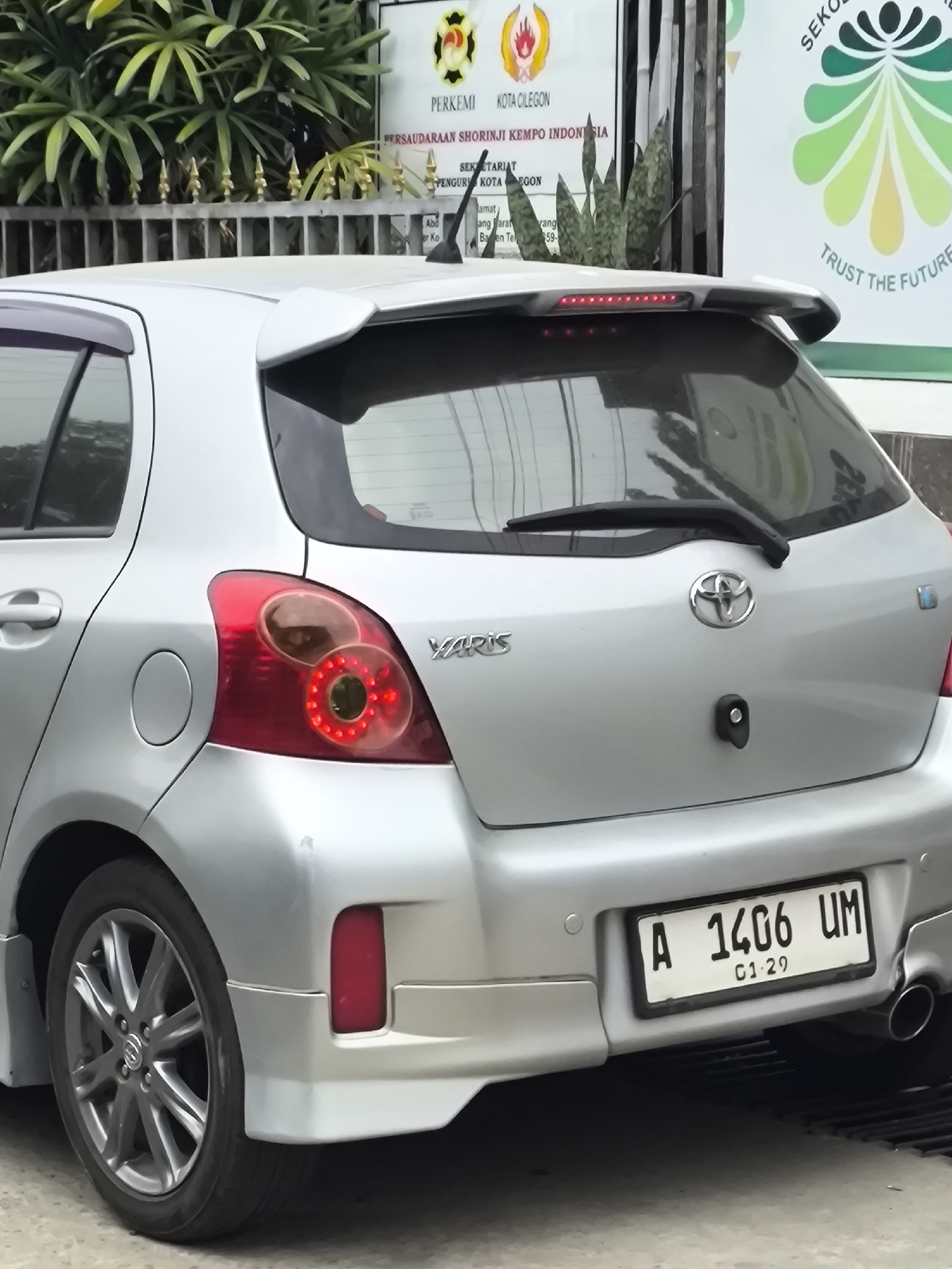 A silver Toyota Yaris is parked on a paved surface. The car is viewed from the rear passenger side, with the right taillight and license plate clearly visible. The license plate reads "A 1406 UM" and a smaller plate below it reads "01 29". A sign with the text "PERKEMI", "KOTA CILEGON", and "PERSAUDARAAN SHORINJI KEMPO INDONESIA" is visible in the background. The sign is green and white with a logo of a stylized flower. The environment appears to be outdoors, with some foliage visible behind the car and fence.