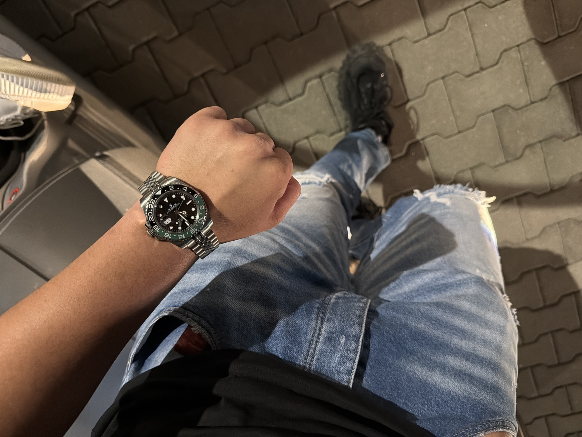 The image provides an overhead perspective of a person seated outdoors, featuring their left arm, wristwatch, and legs. The individual is wearing light blue, heavily ripped jeans and a black garment, likely a t-shirt or hoodie. On their left wrist, a prominent luxury watch is visible. It is a Rolex GMT-Master II, identifiable by its black dial, a metallic Jubilee bracelet, and a distinct bi-color green and black ceramic bezel. Text visible on the watch includes "ROLEX", "GMT-MASTER II", and "SWISS MADE", along with even numbers from 2 to 24 on the bezel.
Two different pairs of shoes can be seen: a light-colored sneaker with a visible ridged sole on the far left, and a dark-colored shoe, possibly a boot or athletic shoe, on the right foot. The ground beneath the person is composed of gray, interlocking concrete pavers, characteristic of a sidewalk, driveway, or parking area. A portion of a metallic grey object, potentially part of a vehicle such as a car door or fender, is visible on the far left side of the frame.
The lighting in the image is dim, suggesting it was taken at night or in very low light conditions, possibly illuminated by artificial sources, which casts subtle shadows. The overall scene conveys a casual and relaxed atmosphere, likely intended to showcase the wristwatch. No specific location cues beyond a paved outdoor area are discernible.