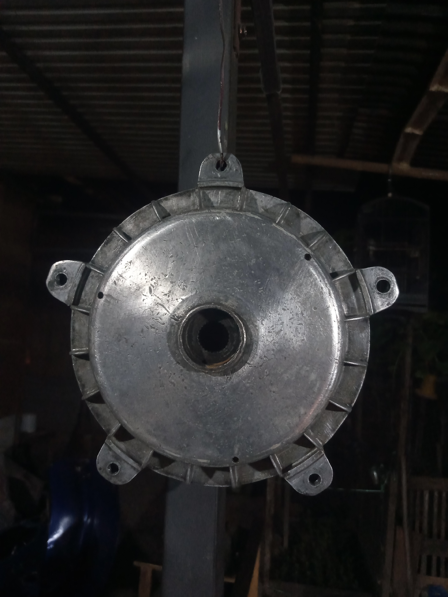 A circular, metallic object, likely a brake drum, hangs suspended by a wire in what appears to be an outdoor workshop or storage area. The object is silver and shows signs of wear, with minor scratches and dirt marks on its surface. It has five mounting points or tabs around its perimeter, each with a circular hole. A central opening reveals splined metal. The background is dimly lit, revealing a corrugated metal roof structure overhead, wooden beams, and some indistinct objects in the shadows, including what could be a birdcage. The scene suggests a utilitarian setting, possibly in a rural or less developed area. The location of Balee Rastong, Indonesia, is not visually discernible from the image itself; it is assumed context provided with the image.