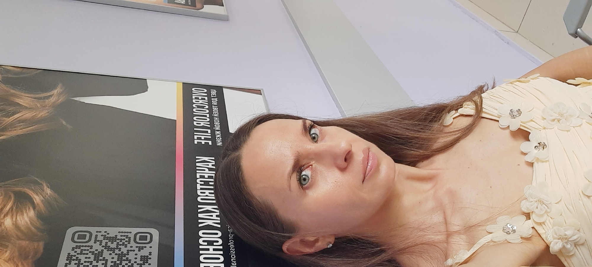 A woman with long brown hair and green eyes is taking a selfie. She is wearing a light yellow dress adorned with fabric flowers and sequins. Behind her, a large poster with text and a QR code is visible, as well as a white wall. The lighting suggests an indoor setting. Based on the text on the poster, which appears to be in Russian and references "Moscow," the image was likely taken in Moscow, Russia. The poster also includes information that could be related to a beauty salon or product, given the text "OVERCOLOR LIFE" and "КАК ОСНОВЕ".