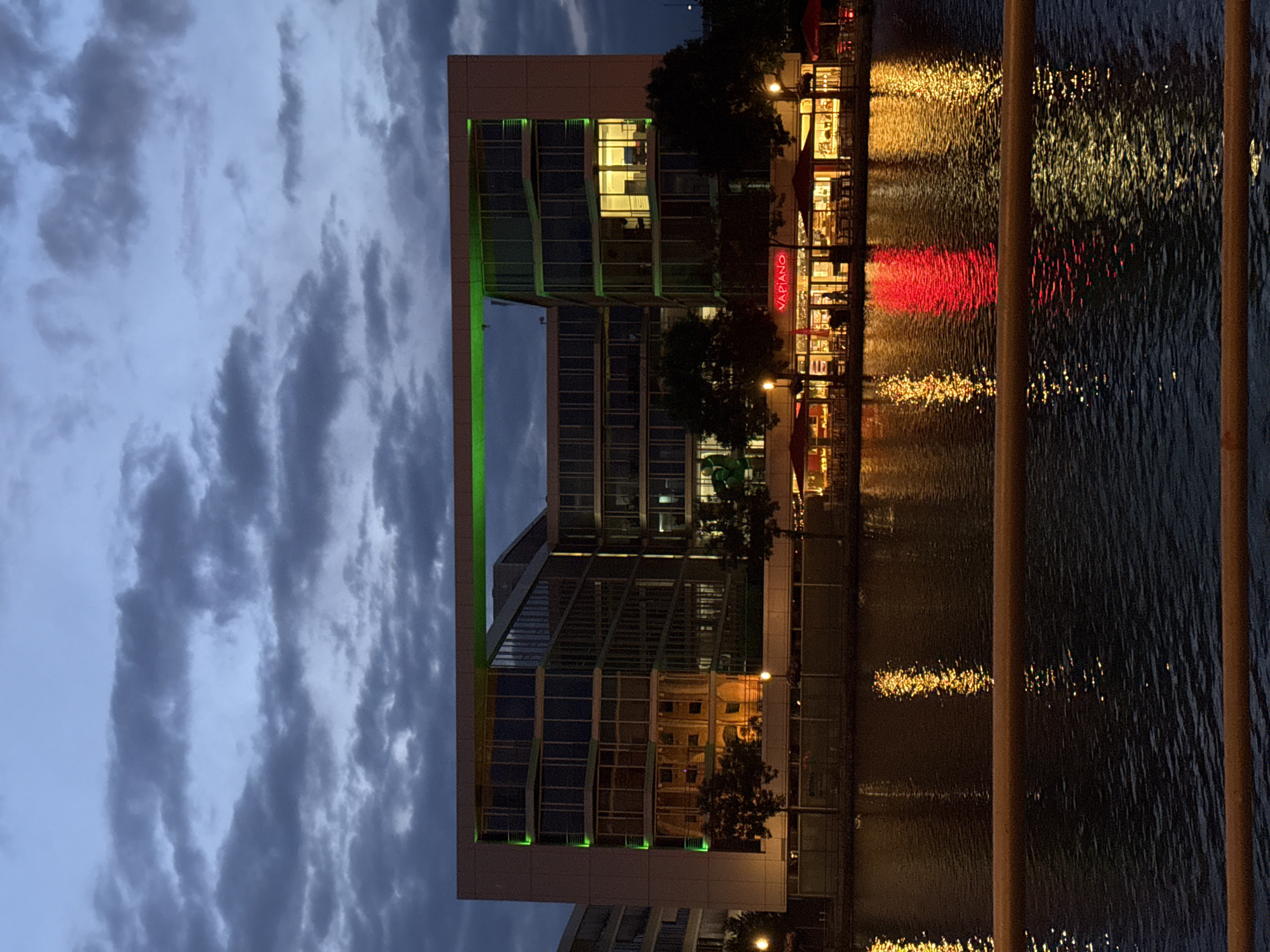 This image depicts the Vapiano restaurant, situated at the edge of the inner harbor in Duisburg, Germany, during twilight. The building, a modern multi-story structure with a large opening in its upper section, is illuminated by green lights along its perimeter and yellow lights from within its windows, indicating occupancy. The restaurant's facade is prominently signed "VAPIANO" in red neon letters. In front of the building, red umbrellas are visible over outdoor seating areas. The scene is reflected in the dark water of the inner harbor, with streaks of red and yellow light shimmering on the surface. The sky above is overcast with dark, textured clouds, contributing to the atmospheric evening mood. A wooden railing runs across the foreground, partially obscuring the view of the water. No individuals are clearly discernible in the image.