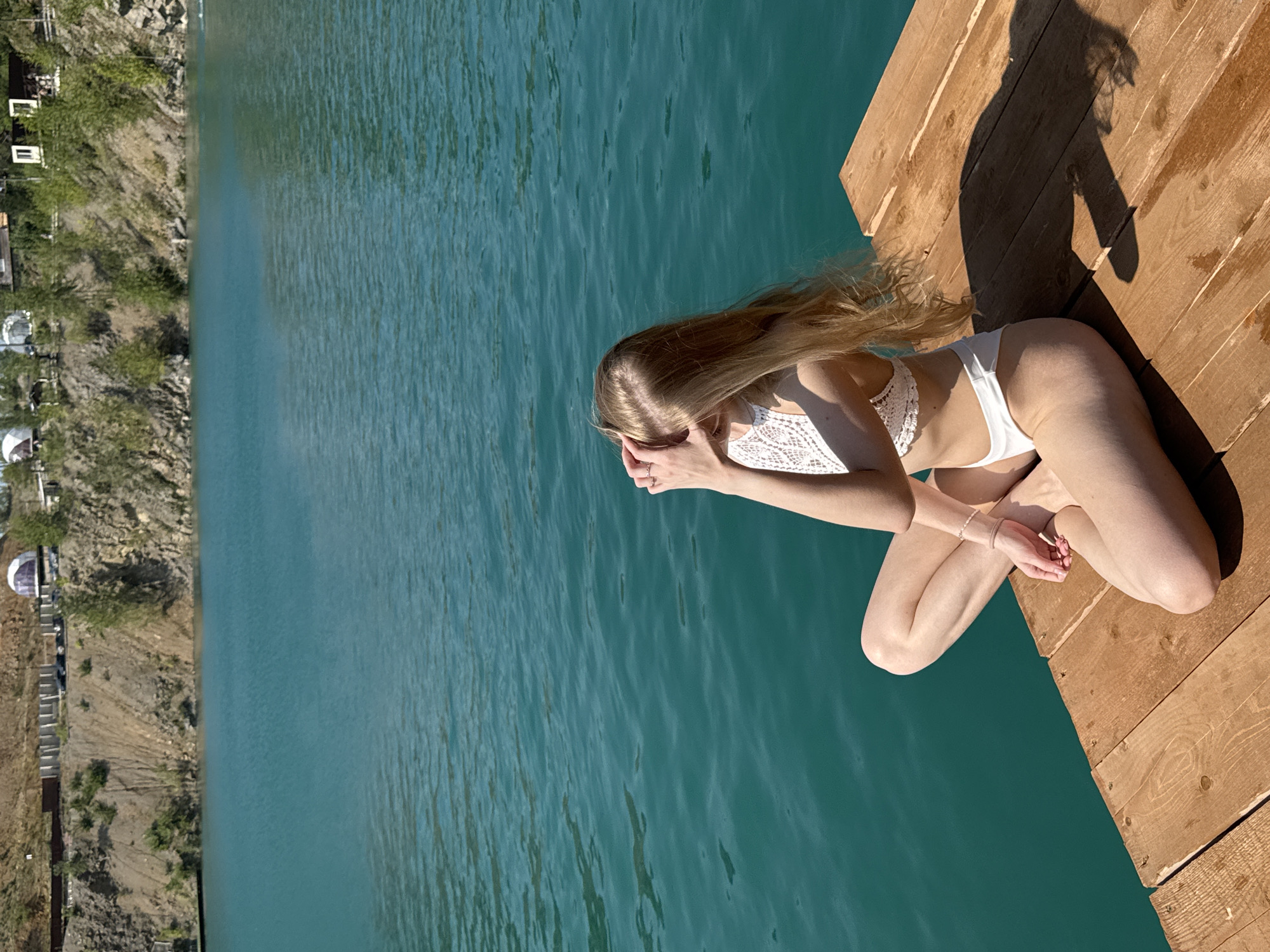 A young woman with long blonde hair, wearing a white bikini, sits cross-legged on a wooden dock. She is facing away from the camera, with her hands over her eyes, as if shielding them from the sun. The dock is situated on the edge of a body of turquoise water, likely the quarry lake at Karyer Izvestnyak, Russia. In the background, a steep, rocky embankment rises from the water, with trees and some structures visible at the top. The sky is clear and blue, indicating a sunny day.