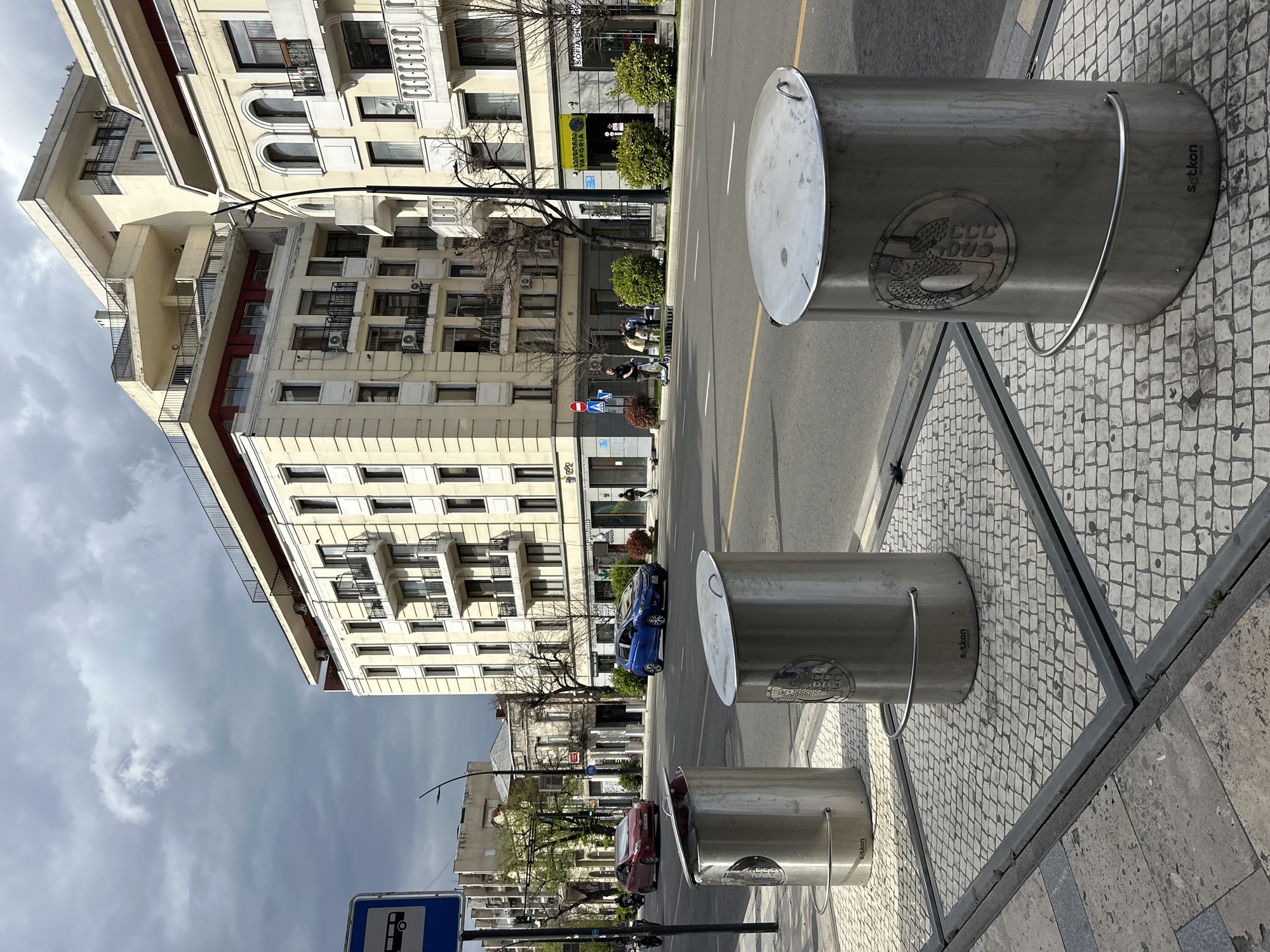 This image shows a street in Tbilisi, Georgia, lined with modern metal trash receptacles. The bins are cylindrical, made of brushed metal, and affixed to a decorative, cobblestone-patterned sidewalk with metal framing. They bear a circular logo. In the background, there's a street with cars (a blue one and a red one are visible) and a multistory beige building with numerous windows and balconies. Trees line the street, some bare of leaves, and the sky is overcast. A bus stop sign is visible in the foreground. Pedestrians are visible in the mid-ground on the sidewalk.