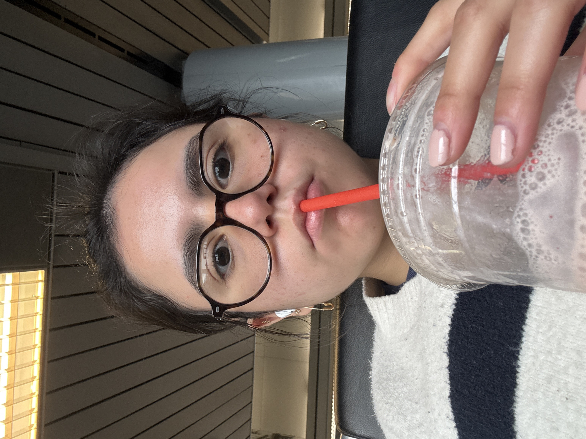 A woman with dark hair, wearing glasses and a white and navy striped sweater, is holding a partially consumed beverage in a clear plastic cup with a red straw. The drink appears to be a smoothie or milkshake. She is indoors in what appears to be an airport waiting area, possibly in Chicago, with linear wall panels and a bank of windows with diffused sunlight in the background. A safety pin is visible in her hair. The seating consists of grey upholstered chairs.
