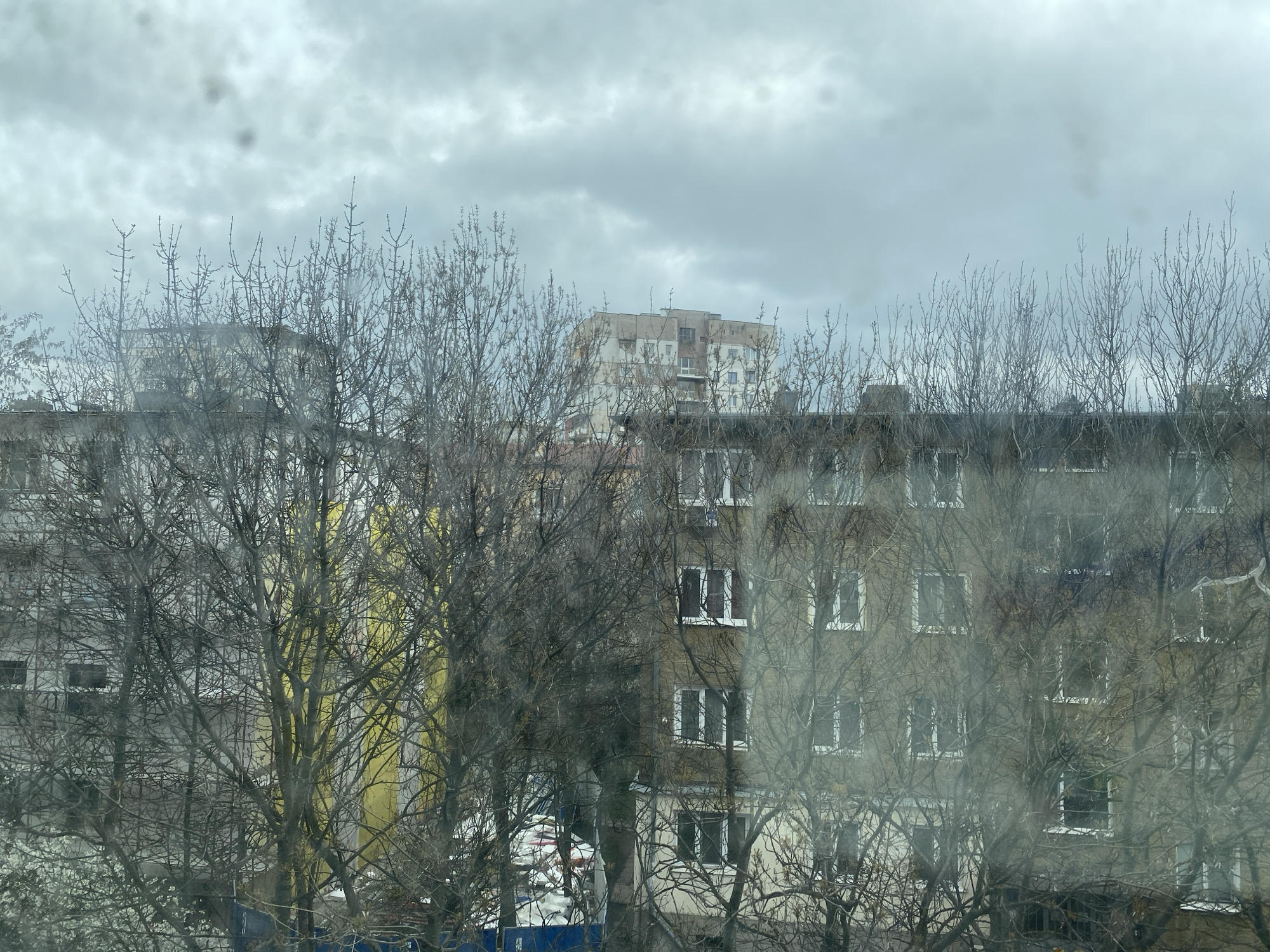 The image shows an overcast urban scene, likely in Sofia, Bulgaria, viewed through a window with water spots. In the foreground, bare trees obscure the view of apartment buildings. The buildings appear to be Soviet-era constructions, characterized by their blocky shape and repetitive window patterns. The sky is uniformly gray. The composition conveys a sense of urban density and a muted atmosphere.