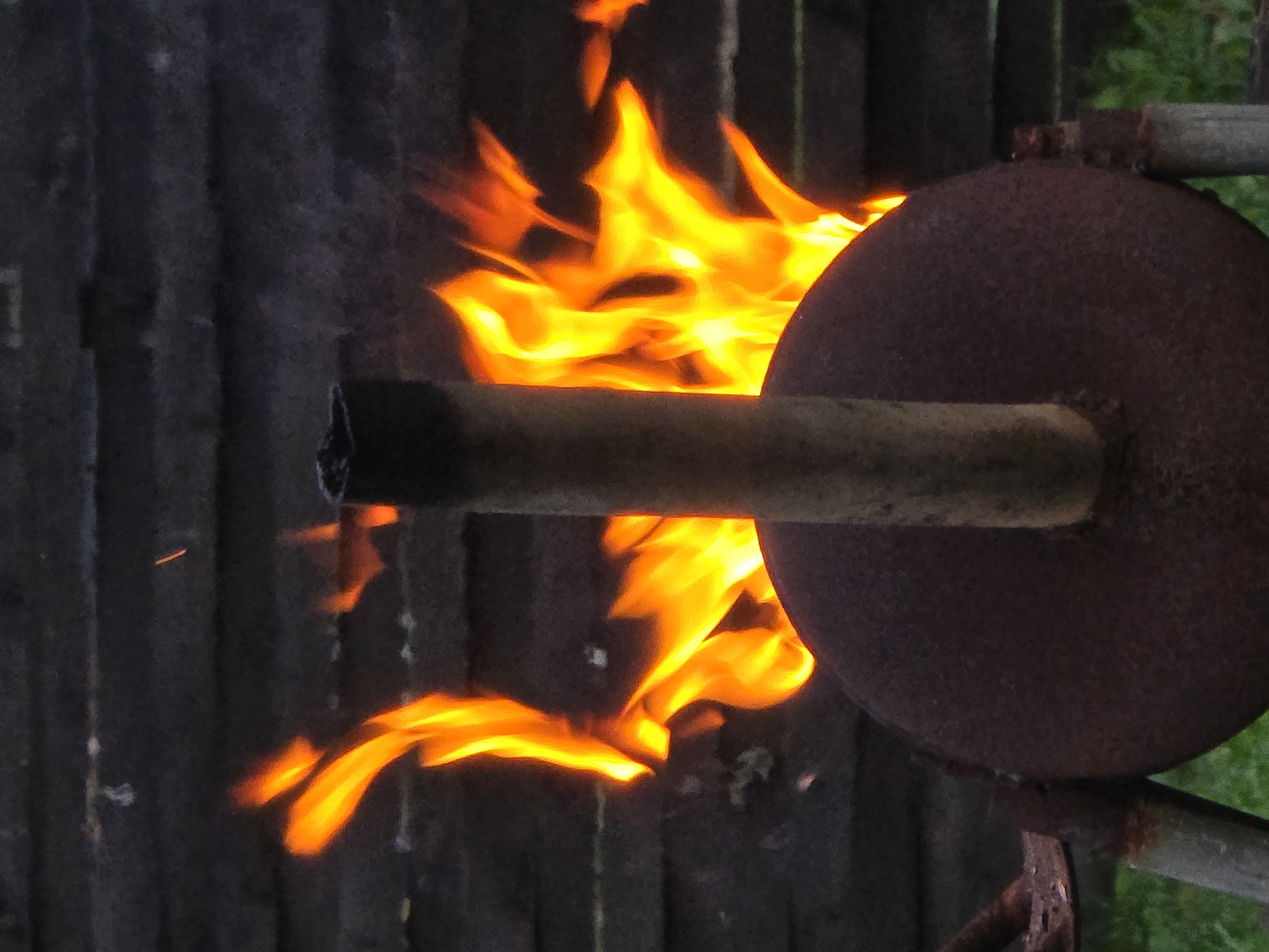 A rusty, circular metal object is positioned in the foreground, with a vertical metal pipe extending upwards from its center. Flames and smoke are visible behind the object, against a dark, wooden fence in the background. The environment appears to be outdoors, with some greenery visible at the bottom of the frame. The scene suggests a fire is burning, possibly for cooking or heating, in an outdoor setting that could be in a rural or suburban area like Ермолаево, Russia. No people are present in the image.