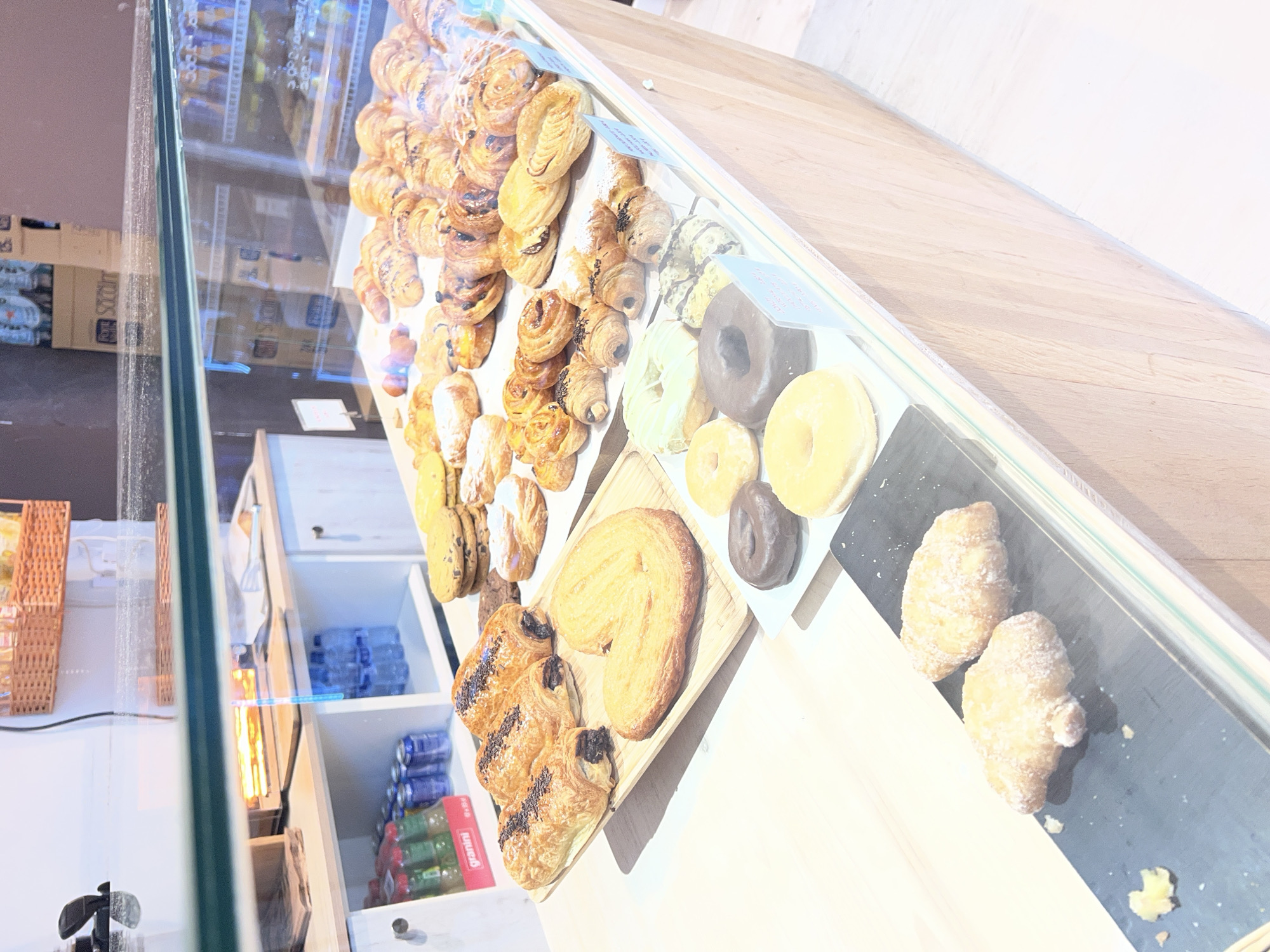 This media file captures a bright, slightly overexposed interior view of a bakery or pastry shop display counter, presented from an angled, top-down perspective. The primary subjects are a diverse array of freshly baked goods arranged within a glass display case and on adjacent wooden surfaces.
Prominently featured are numerous golden-brown pastries, including various types of croissants (plain, chocolate-filled, and possibly pain aux raisins with visible dark fruit), palmier cookies, and powdered sugar-dusted pastries resembling small croissants or twists. A section also showcases several donuts with different toppings, such as chocolate glazed and plain sugar glazed, alongside what appears to be large, flat cookies or similar items. Many of the pastries are displayed on wooden trays or directly on the counter, with some appearing to be on light-colored tags or labels, though the text on these tags is unreadable due to blurriness and distance.
The setting is a retail environment, indicated by the display case and the background reflections. Through the glass, reflections show shelves stocked with various products, including bottled water, canned drinks, and cardboard boxes. Text is partially visible on some reflected items, with "Sucorn" seen on a box and "Granini" on a package of juices. Below the main display, glimpses of white shelves holding more bottled drinks are visible.
No people are present in the image. The lighting is artificial and bright, suggesting an indoor setting during operating hours. The overall impression is that of an inviting selection of baked goods ready for purchase in a clean and well-stocked establishment, likely a cafe or a bakery in Barcelona, Spain.
