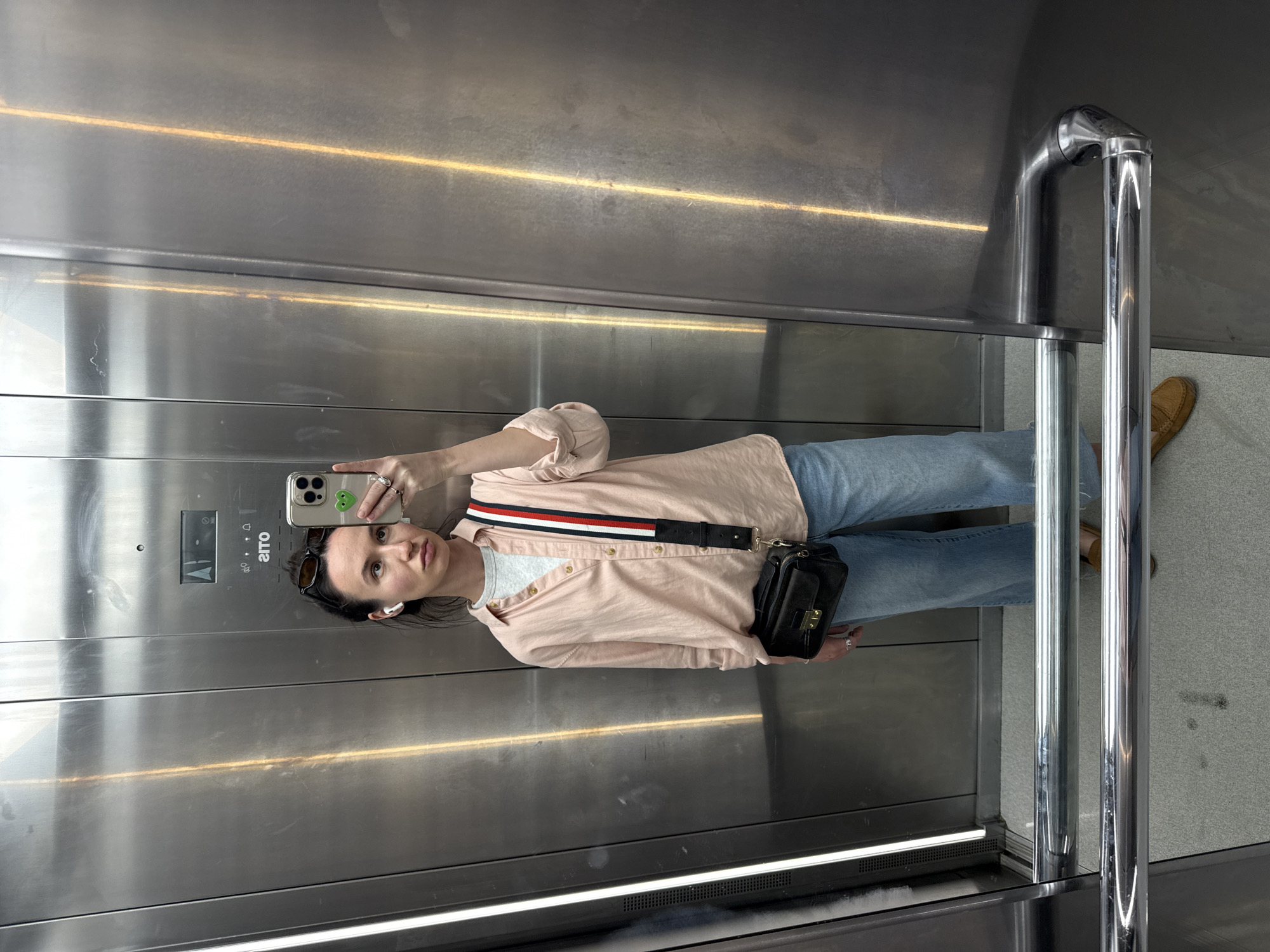 An individual is captured in a mirror selfie taken inside an elevator. The person, a woman, stands facing the reflective metallic surface of the elevator, holding a smartphone with a green heart sticker on its case to capture her reflection. She has dark hair pulled back, sunglasses resting on her head, and white wireless earbuds. She is dressed in a light pink long-sleeved button-up shirt worn open over a light grey t-shirt, blue jeans, and tan loafers. A black cross-body bag with a distinctive red, white, and blue striped strap hangs across her torso.
The setting is an elevator interior, characterized by brushed metallic walls and a chrome handrail visible on the right. An elevator control panel is reflected on the left side of the image, showing buttons and a display with the mirrored brand name "OTIS" clearly visible. The floor is light grey. The internal elevator lighting creates horizontal reflections across the metallic walls. The overall scene suggests an indoor, modern environment, possibly in Tel-Aviv, Israel.