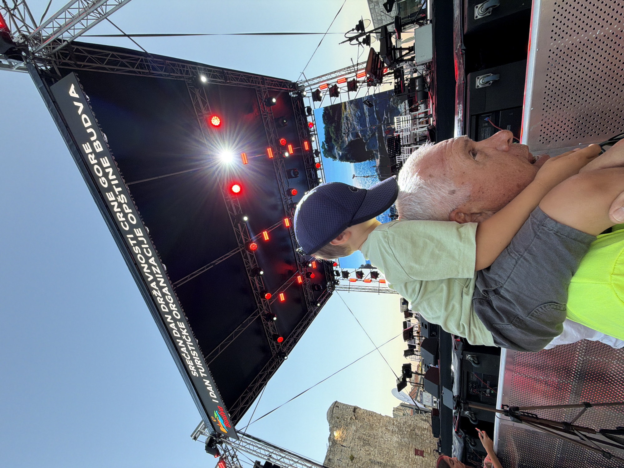 An outdoor event is taking place in front of a historical stone structure, likely in Budva, Montenegro, given the signage on the stage. A man with gray hair is seen from the side, with a young boy sitting on his shoulders. The boy is wearing a light green t-shirt and a blue baseball cap. The man is wearing a bright yellow shirt. They are looking towards a large stage setup, which includes a prominent screen displaying a scenic view of a rocky coastline. The stage is lit with red and white lights, and there are speakers and other audio equipment visible. The sky is clear and blue, suggesting it is daytime. In the background, a section of an old stone wall or building stands to the left of the stage. There are also metal barricades in the foreground, suggesting a controlled access area for the event.