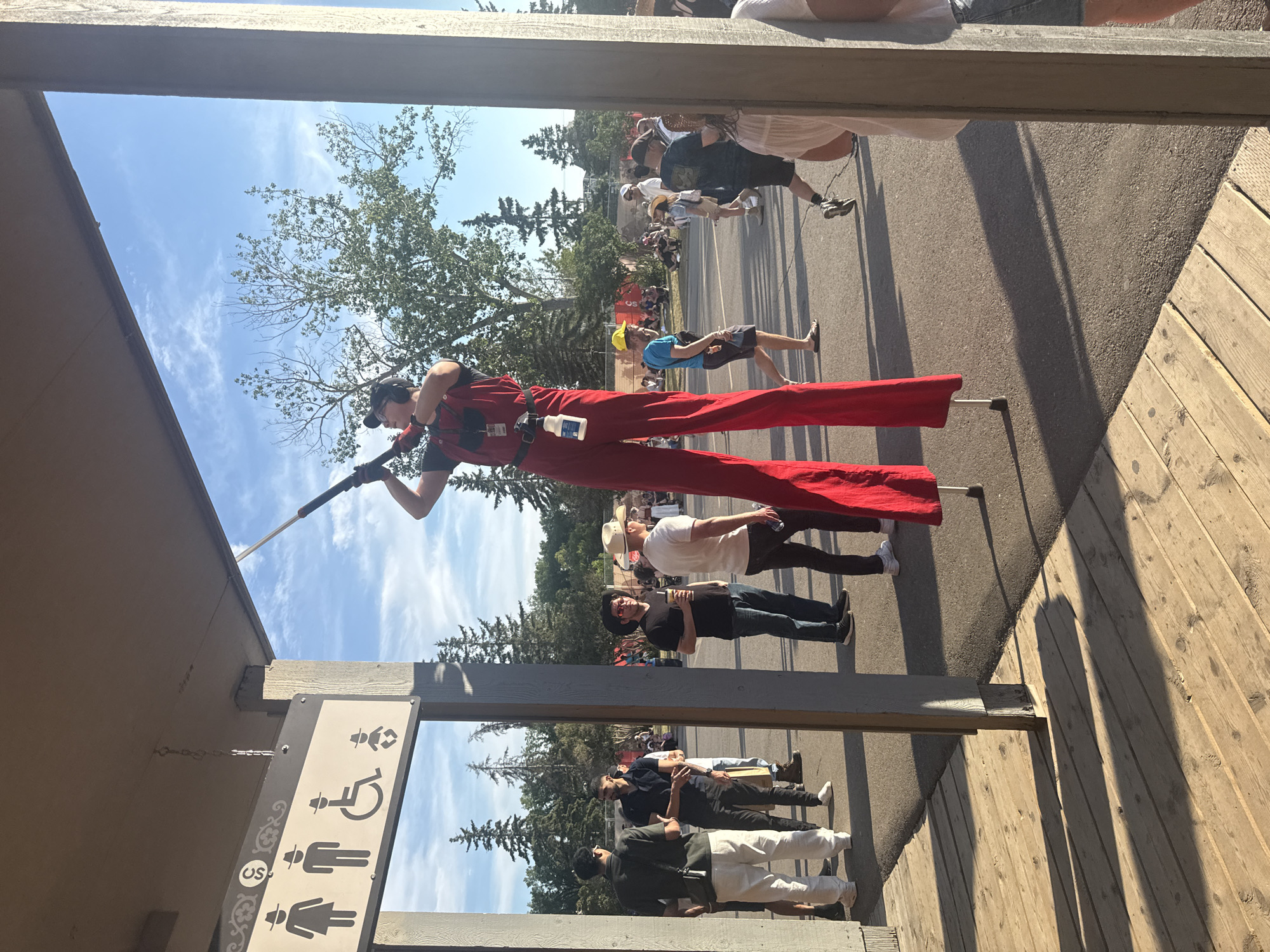 The image shows a bright, outdoor daytime scene, likely at an event or festival, with a clear blue sky and scattered clouds. In the foreground, a wooden deck with prominent shadows from the setting sun is visible. To the left, a sign with restroom symbols for men, women, disabled individuals, and infants is attached to a wooden structure.
The main subject is a performer on stilts, dressed in a red jumpsuit and black headphones, holding a long, thin pole. The performer is positioned in the center of the frame and appears to be interacting with something overhead.
In the background, a crowd of people are milling about. Many are casually dressed, with some wearing hats that suggest a Western or festival atmosphere. The ground is paved, and trees are visible in the distance. The overall impression is of a lively, public gathering. Given the signage mentioning "US" which could stand for United States, and the style of the event, it is possible this is a border city or a festival that draws international visitors. However, without further context, it's difficult to pinpoint the exact location within Calgary, Canada.
