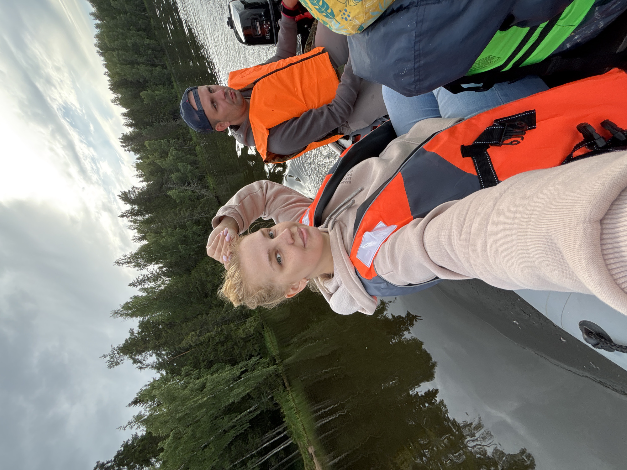 The image shows two people, a woman and a man, in an inflatable boat on a body of water. The woman is in the foreground, taking a selfie. She has blonde hair pulled back and is wearing a light-colored hoodie and an orange life vest. The man is in the background, wearing a baseball cap and an orange life vest, and appears to be operating the boat. The boat is moving, as indicated by the ripples on the water and the spray behind it. The background consists of a dense forest lining the shore of the lake. The sky is overcast with some patches of sunlight. The overall impression is a recreational boat trip on a lake, possibly in a rural or natural area. The location is identified as Anteplevo (Okhotinskoe S/P), Russia.