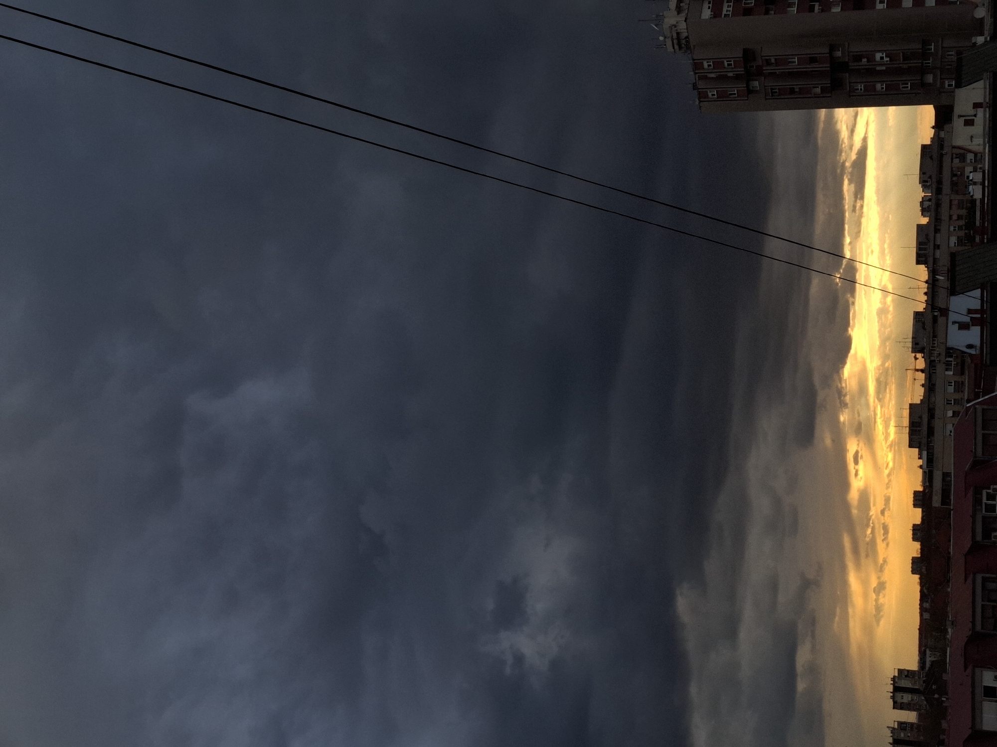 The image displays a dramatic urban skyscape, characterized by a stark contrast between dark, heavy clouds and a brilliant golden-orange horizon. The scene is presented with the horizon running vertically along the right side of the frame, suggesting the original photograph might have been taken in portrait orientation or rotated.
On the left and upper parts of the image, the sky is dominated by dense, deep grey and blue-grey clouds, indicating overcast or potentially stormy weather. These clouds show varying textures and shades, with some areas appearing almost black.
Towards the right side, the clouds transition dramatically into a luminous band of golden and orange light, strongly suggesting either a sunset or a sunrise. This intense light illuminates the edges of the clouds, creating a vibrant glow.
Two dark, thin power lines or cables stretch diagonally across the upper left portion of the dark sky. Along the right edge of the image, silhouetted urban buildings are visible against the bright, glowing sky. These appear to be multi-story residential or commercial structures, with one taller building in the upper right showing discernible windows and architectural details. The buildings collectively suggest an urban or suburban setting.
There are no people, animals, or specific activities depicted in the image. No visible text is present. The notable details include the time of day, which is clearly dusk or dawn due to the intense golden light, and the cloudy weather conditions.