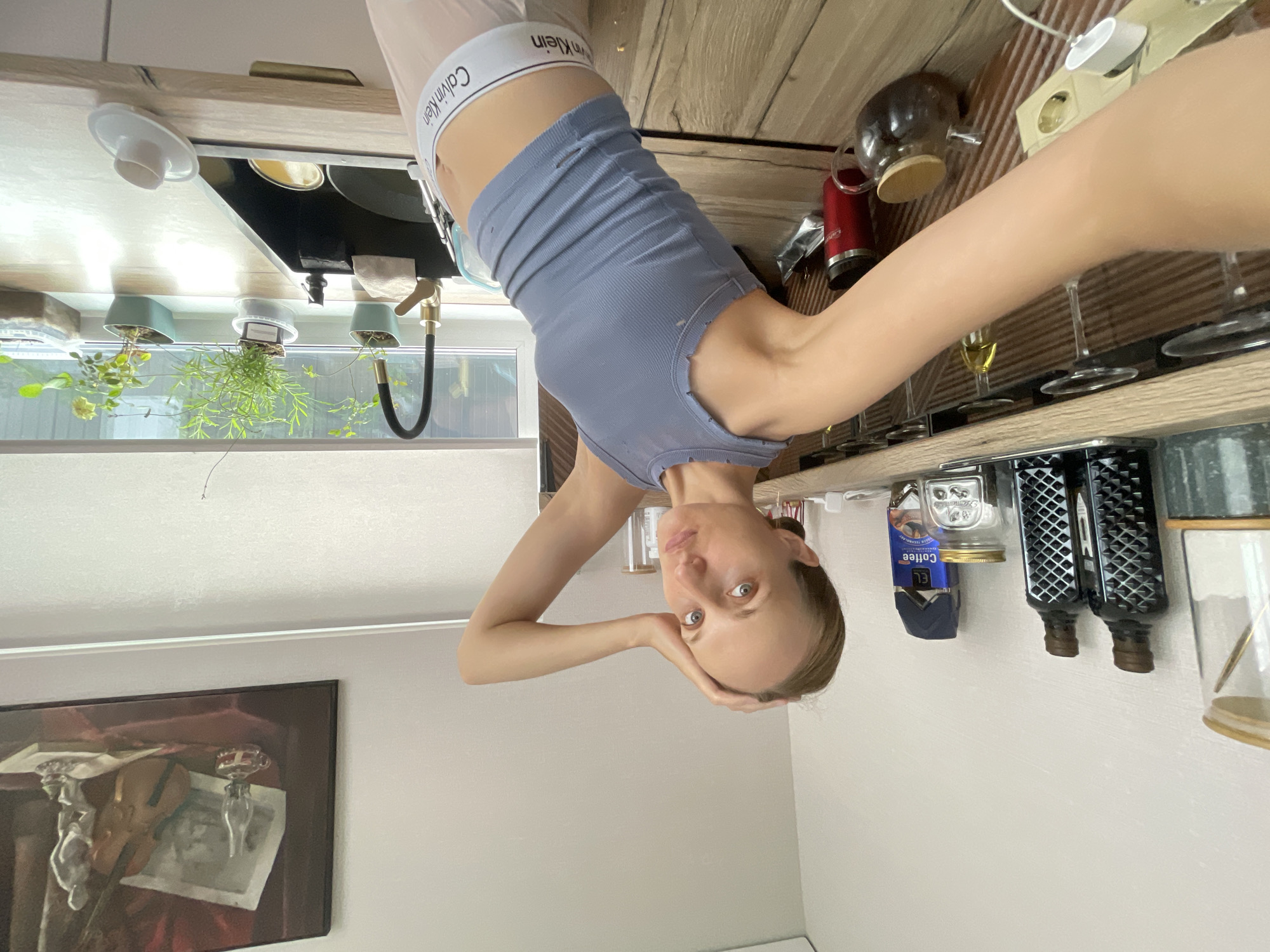 A woman is taking a selfie in a kitchen. She is wearing a gray tank top and light pink underwear. Her left arm is raised, and her hand is touching her head. The kitchen has wooden countertops and cabinets. There is a sink with a faucet and plants in the background. On a shelf above the counter, there are several bottles, a box of coffee, and a glass jar. A painting hangs on the wall above the counter.