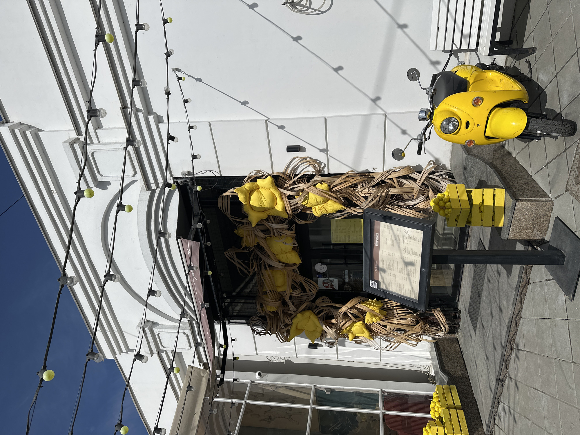 The image shows the exterior of a building with architectural details, possibly in Yekaterinburg, Russia (based on the menu text on display). A bright yellow scooter is parked on the sidewalk. The building's entrance is decorated with a combination of woven straw elements and yellow flower-shaped cushions. A menu board is displayed on a black stand near the entrance. Strings of lights hang along the building's facade. Several yellow crates filled with round objects (possibly lemons or other yellow fruit) are stacked near the base of the building. The day is sunny, with a blue sky visible.