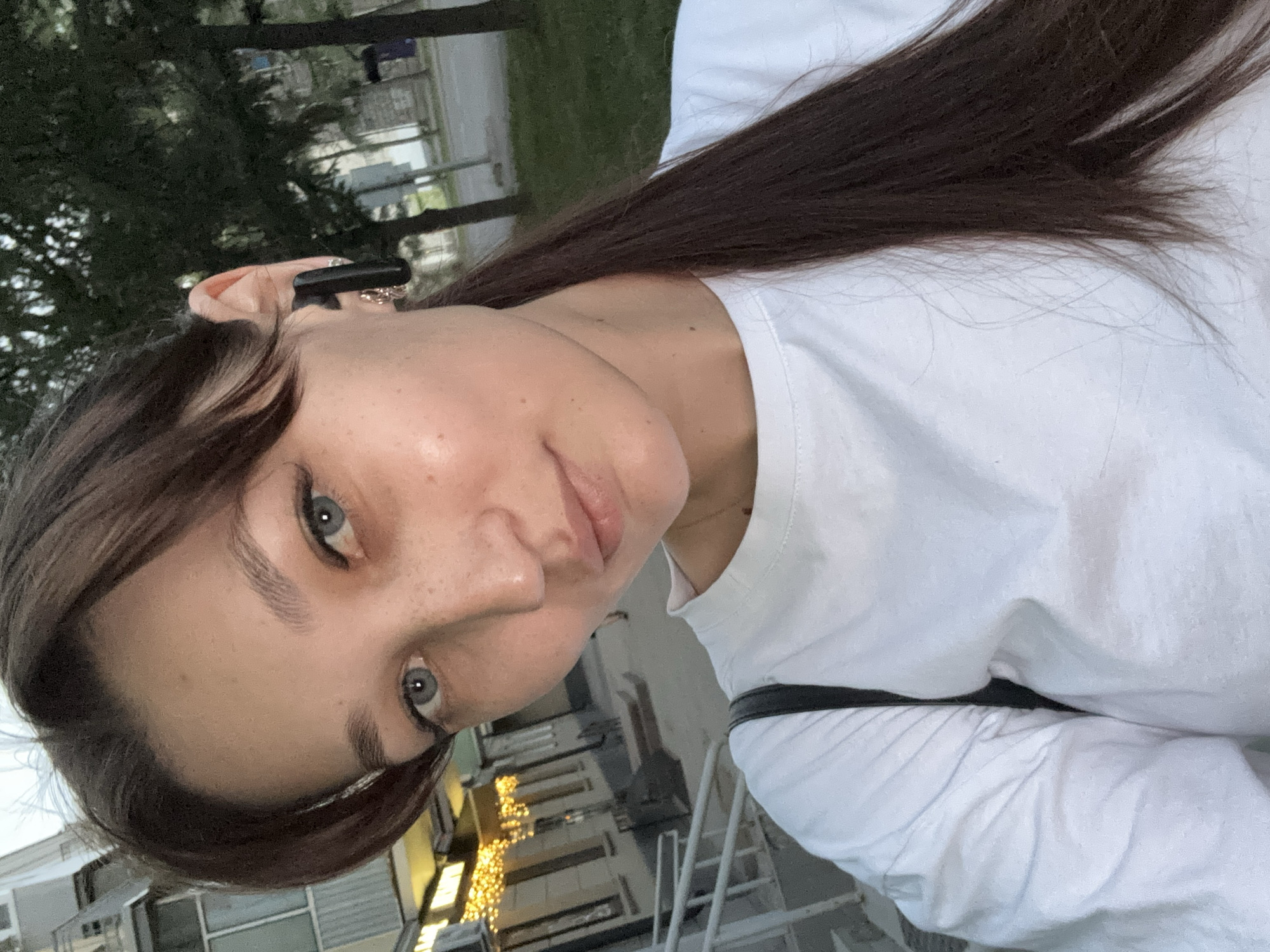 A selfie-style, medium-shot image shows a young woman with dark brown hair pulled back into a ponytail. She has fair skin with freckles and is wearing a white t-shirt. A black wireless earbud is in her right ear. She is outdoors, with a building facade featuring yellow string lights visible to her left and trees and a street in the background. The location is Novokuznetsk, Russia.