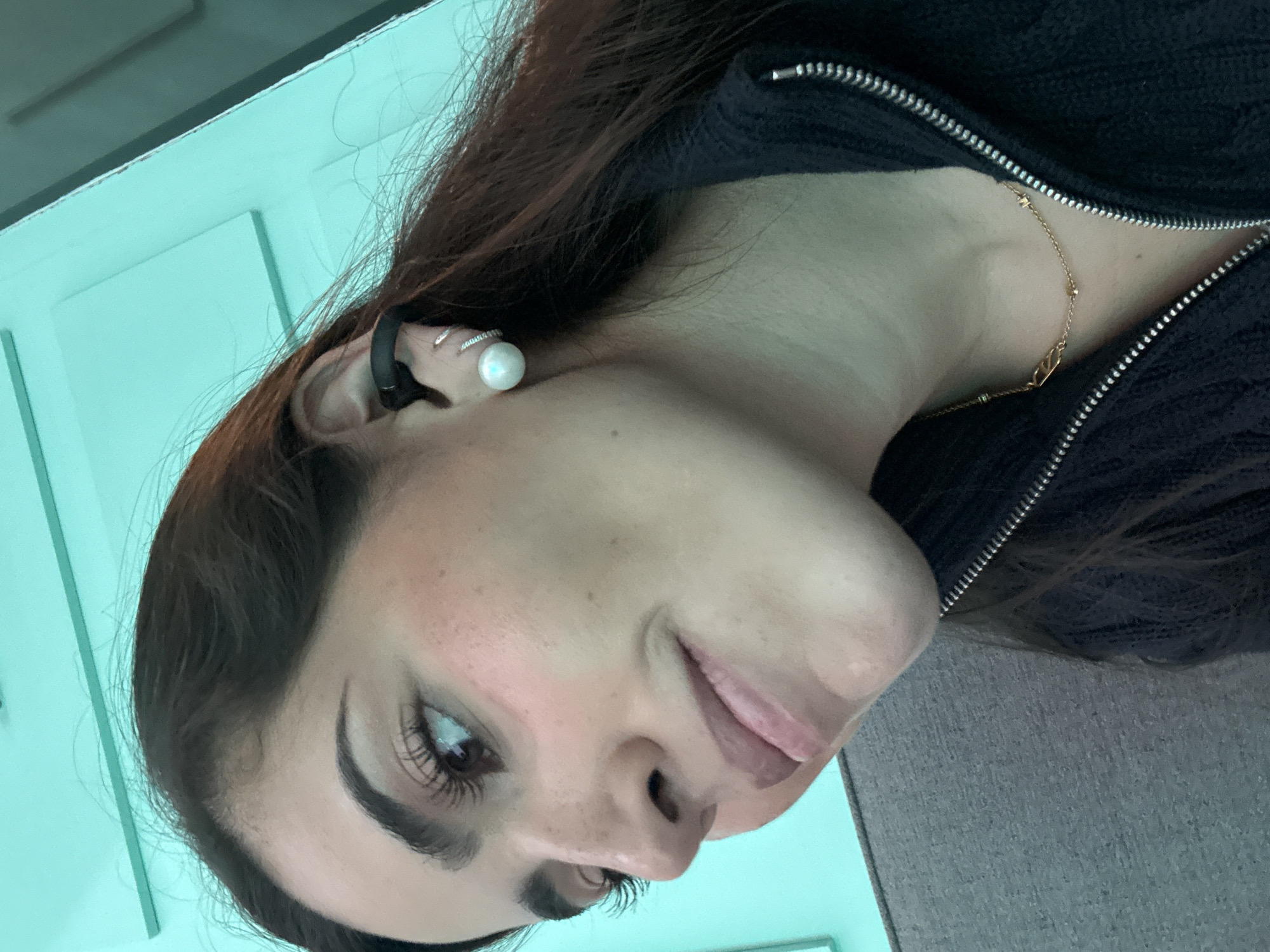 A woman with dark hair is visible from the chest up. She is wearing a black zippered top and a gold necklace. She has a pearl earring in her left ear, along with a black electronic device that wraps around her ear. She is facing toward the left of the frame with her head tilted. The background is a pale blue-green color. There is no information available about the location within San Bernardino Tlaxcalancingo, Mexico.